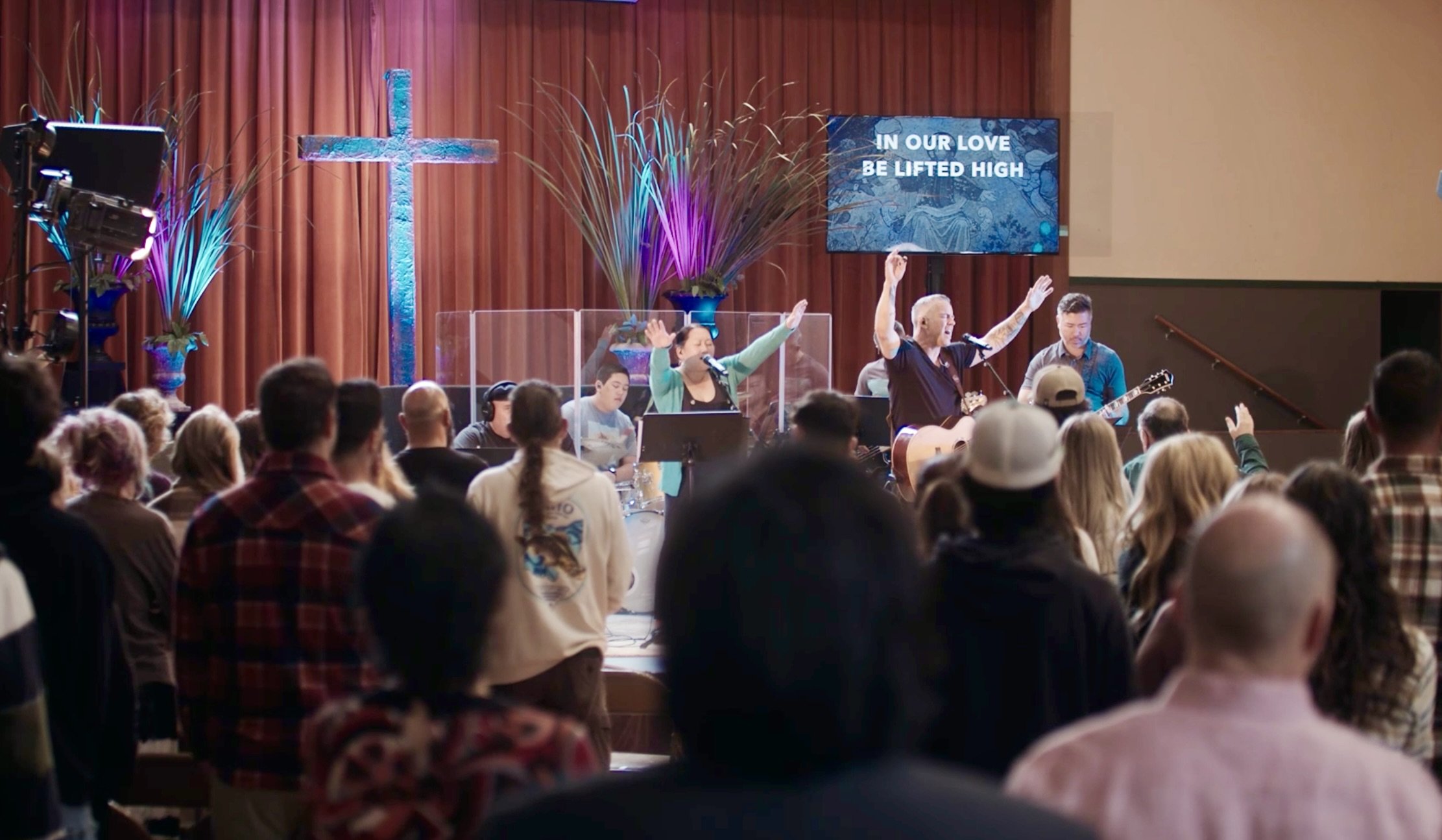 A church service or worship gathering with a band performing on stage in front of a congregation. The stage has a cross and floral decorations. Some people are singing or playing musical instruments, with a screen displaying the lyrics, 'IN OUR LOVE BE LIFTED HIGH'.