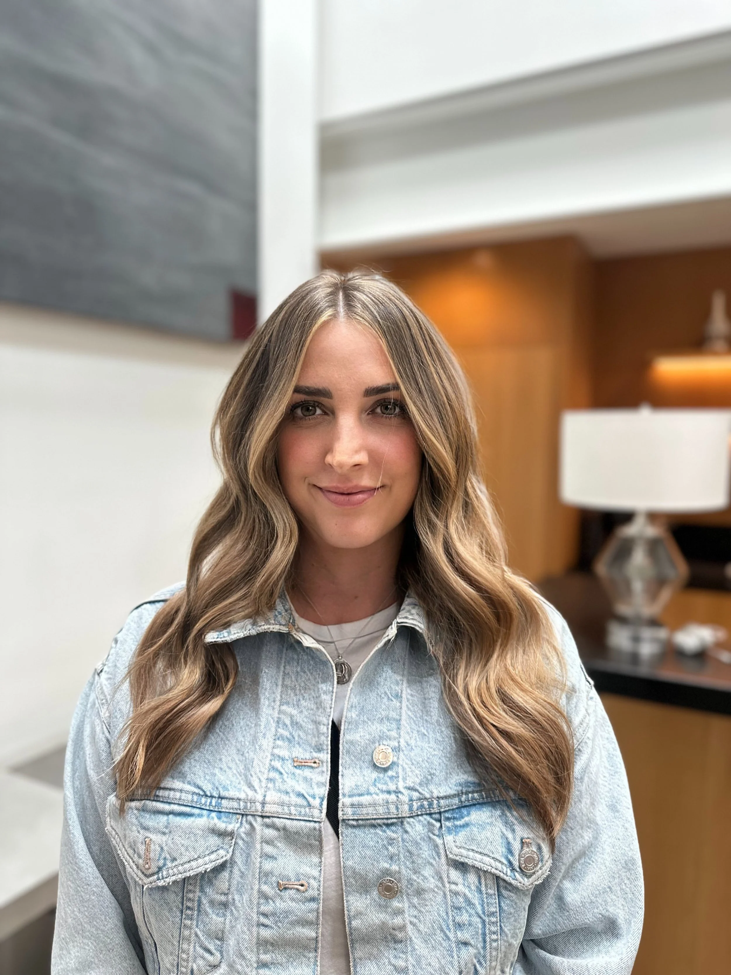 A young woman with long, wavy hair smiling in an indoor setting, wearing a light denim jacket and a white top.