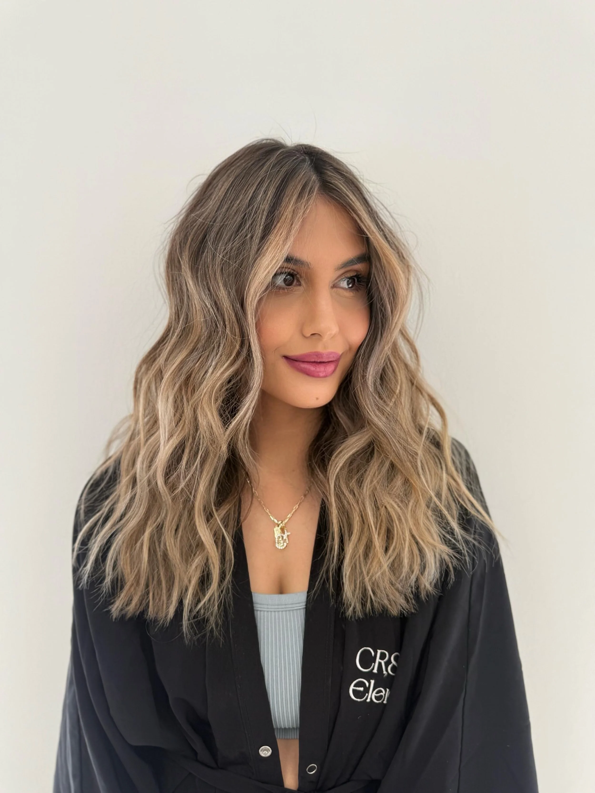 A woman with long, wavy, blonde hair and pink lipstick is smiling slightly and looking to her left. She is wearing a black jacket with white text and a gray top underneath, and a gold necklace with a pendant.