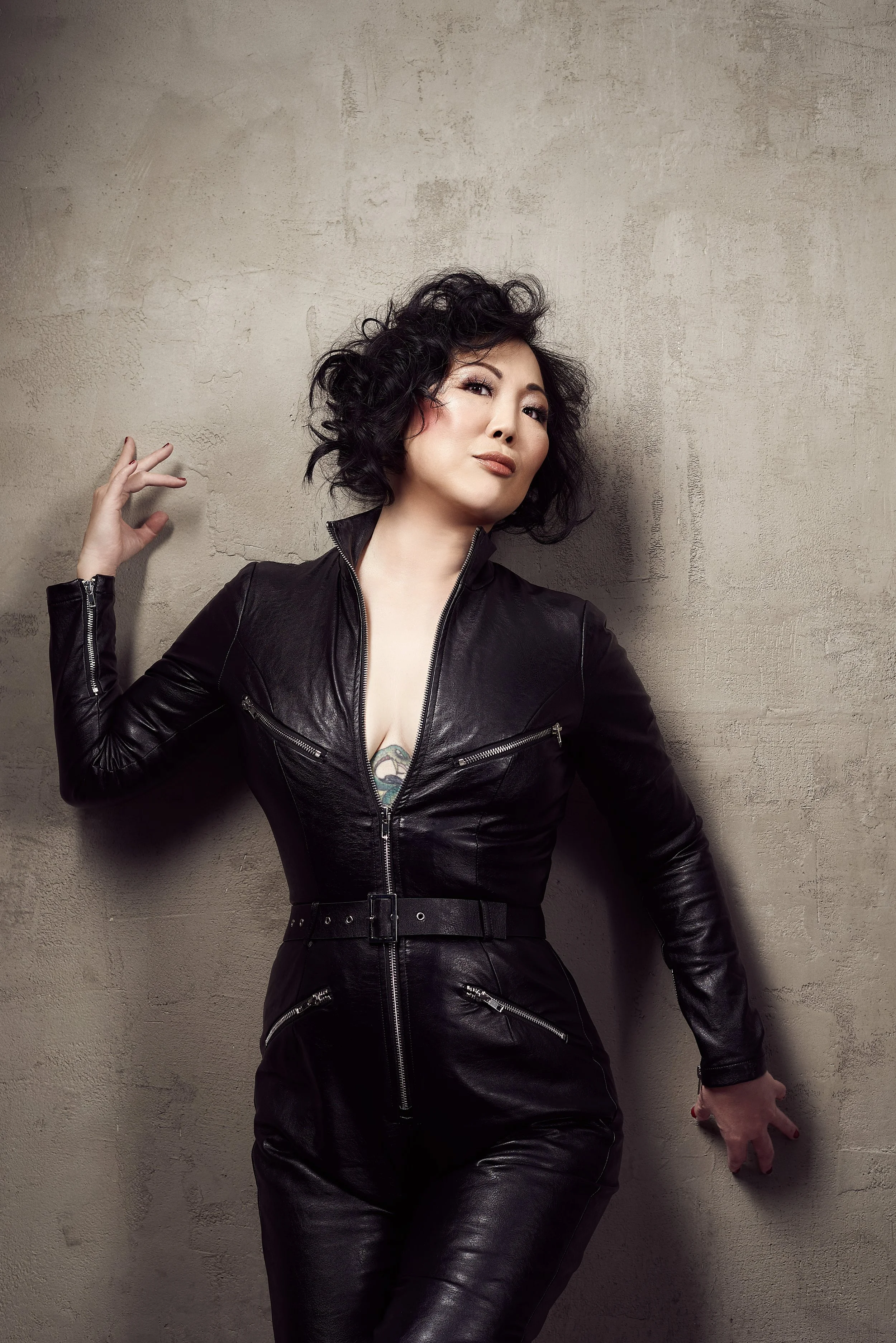 A woman with short, curly black hair wearing a black leather jumpsuit standing against a textured beige wall.