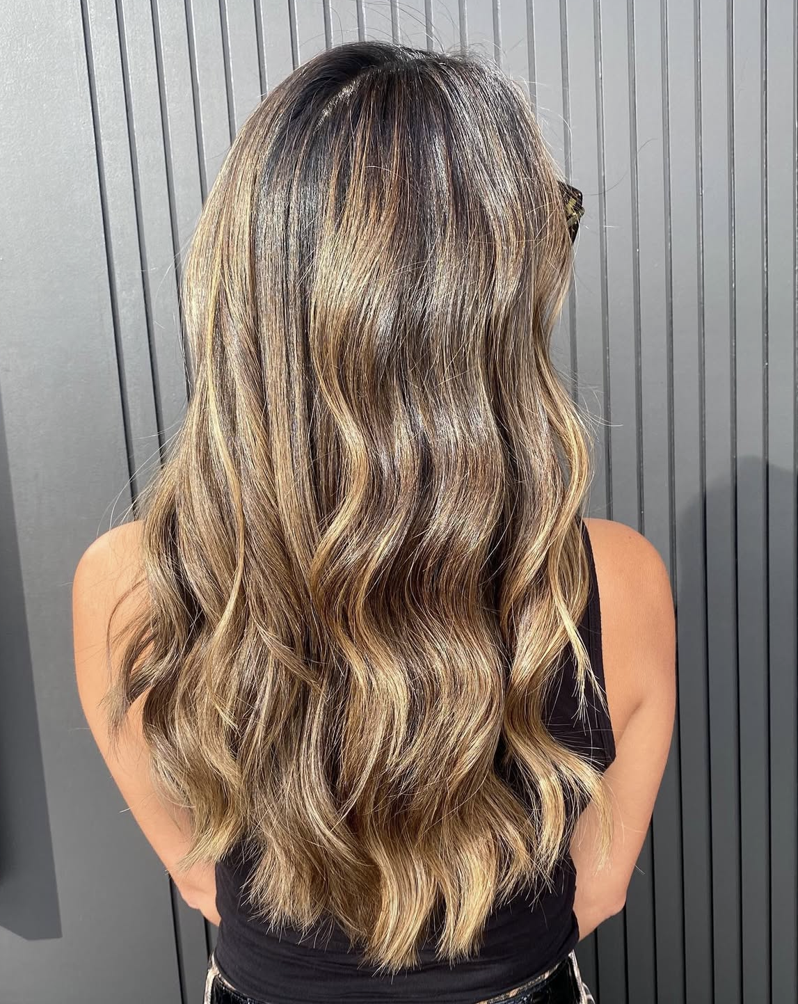 Back view of a woman with wavy, highlighted hair in shades of brown and blonde, standing in front of a grey, paneled wall.