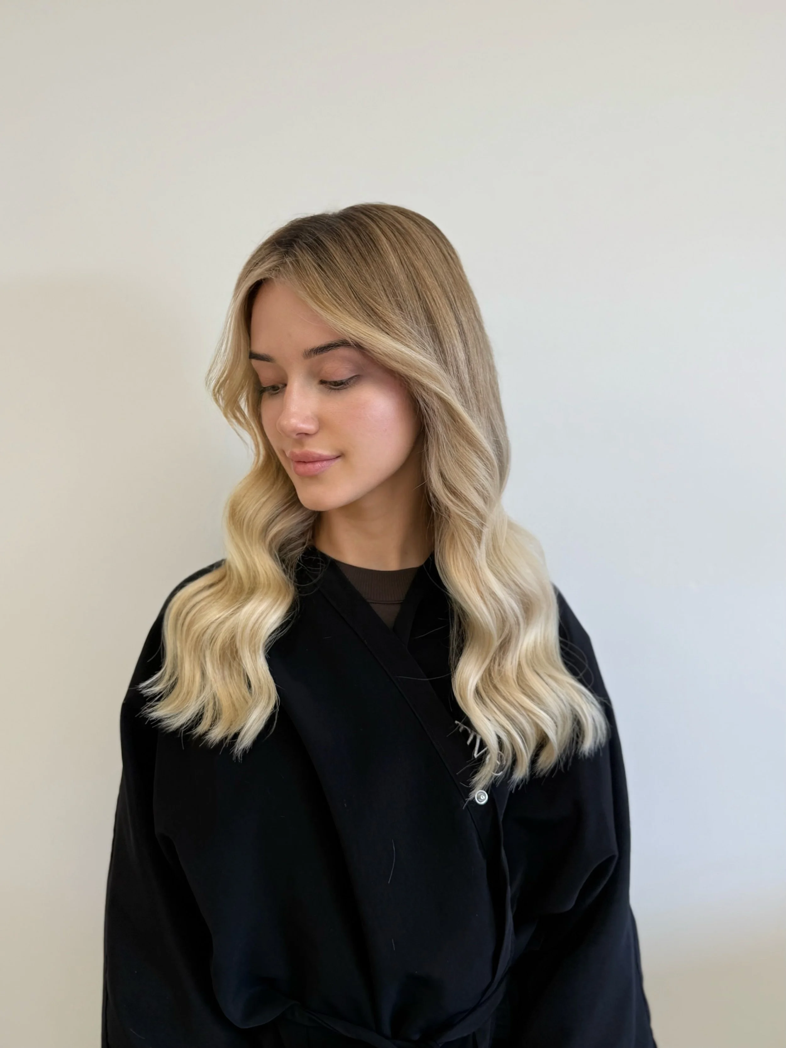 A woman with long blonde wavy hair looking down, wearing a black top, standing against a plain white wall.
