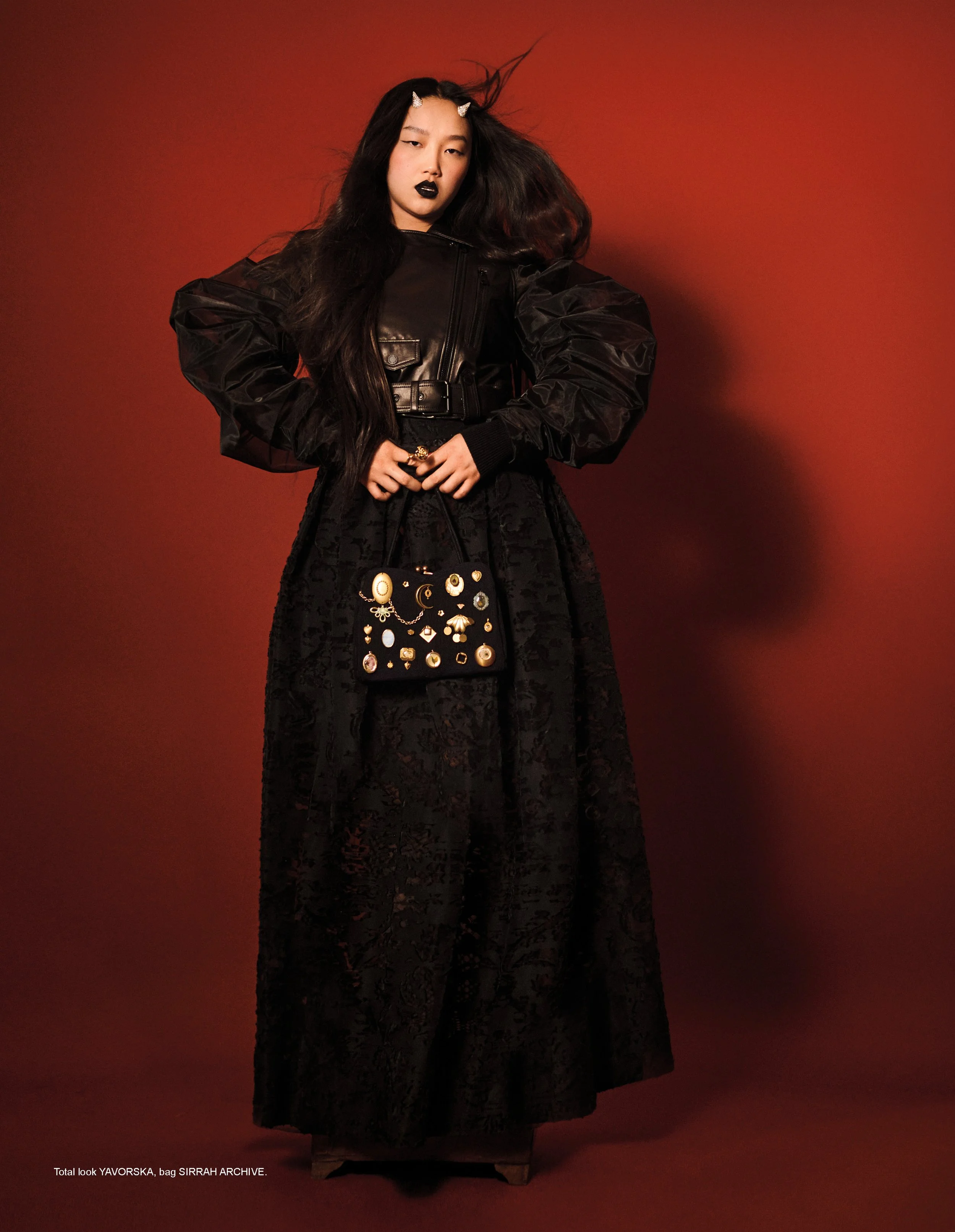 A woman with long black hair and dark lipstick stands against a red background, wearing a black leather jacket, a long black lace skirt, and holding a small black handbag decorated with gold and jewel embellishments.