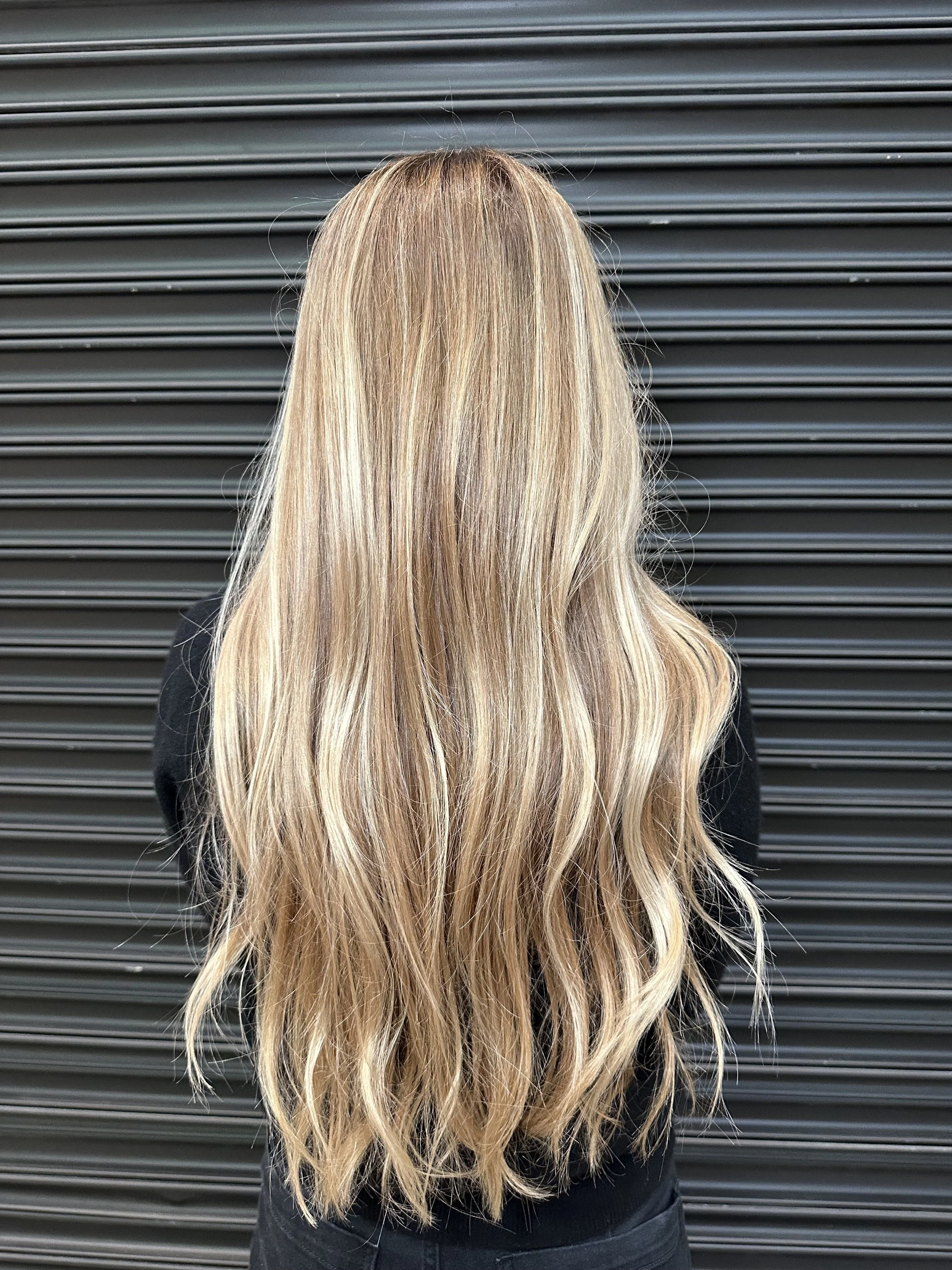 Back view of a woman with long, wavy blonde hair standing in front of a black corrugated metal wall.