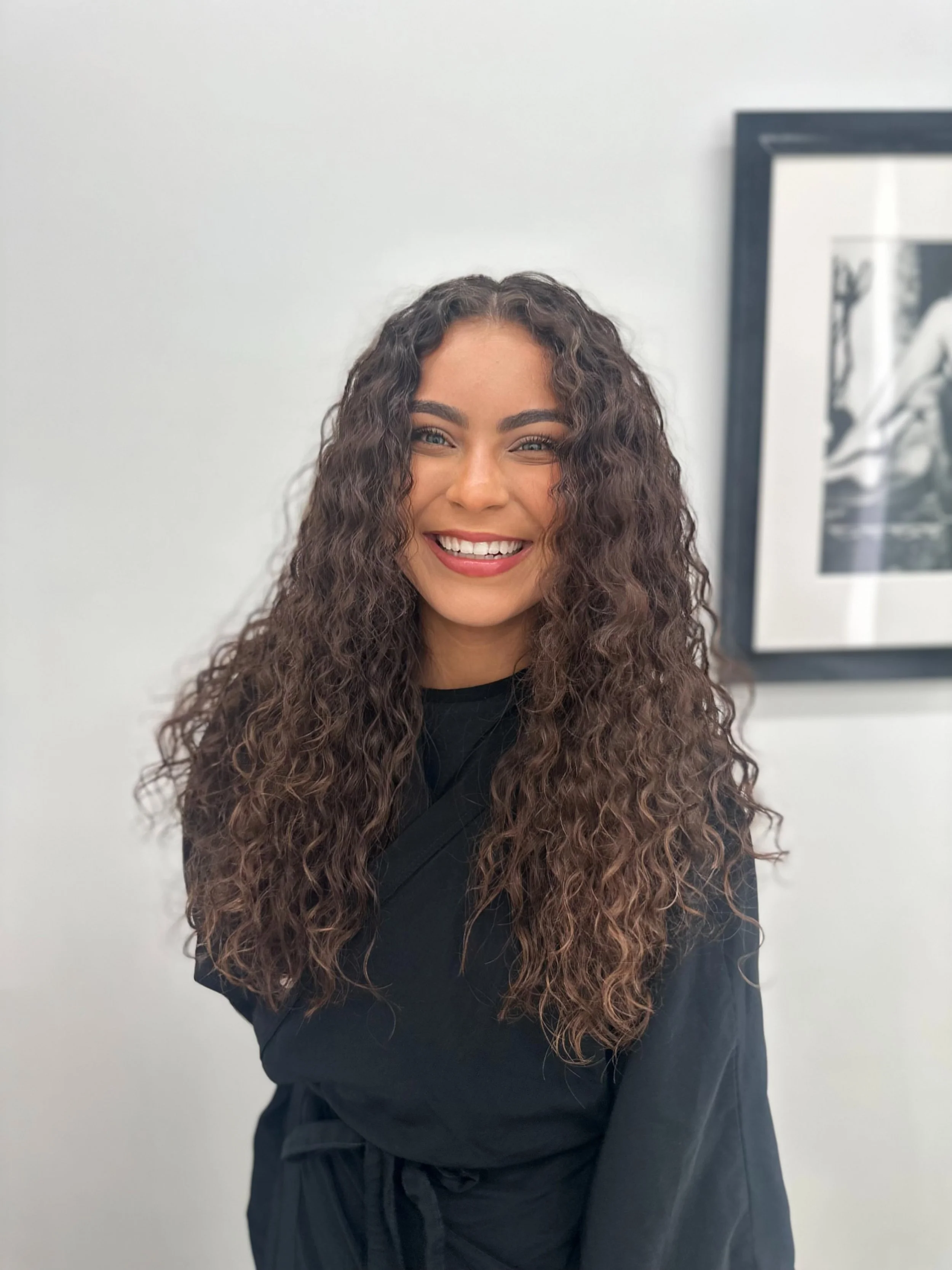 Bright smiling woman with long curly hair standing in front of a white wall and framed artwork.