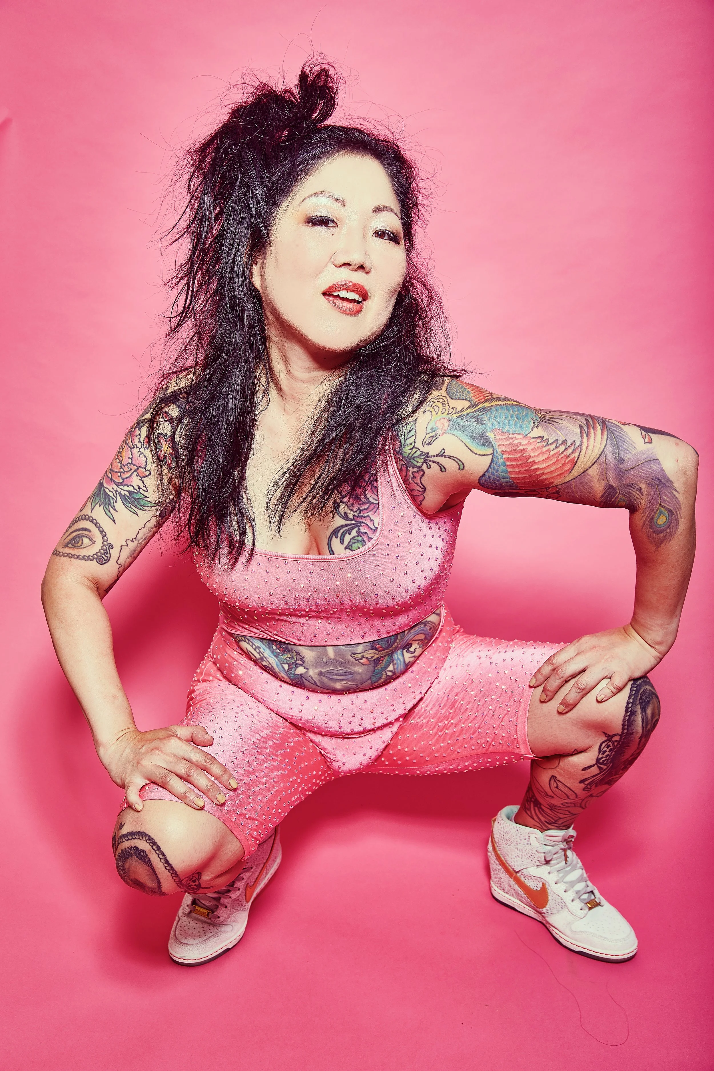 A woman with dark, messy hair poses in a squatting position against a pink background. She wears a pink, studded athletic outfit and white Nike sneakers, and has numerous colorful tattoos on her arms, torso, and legs.