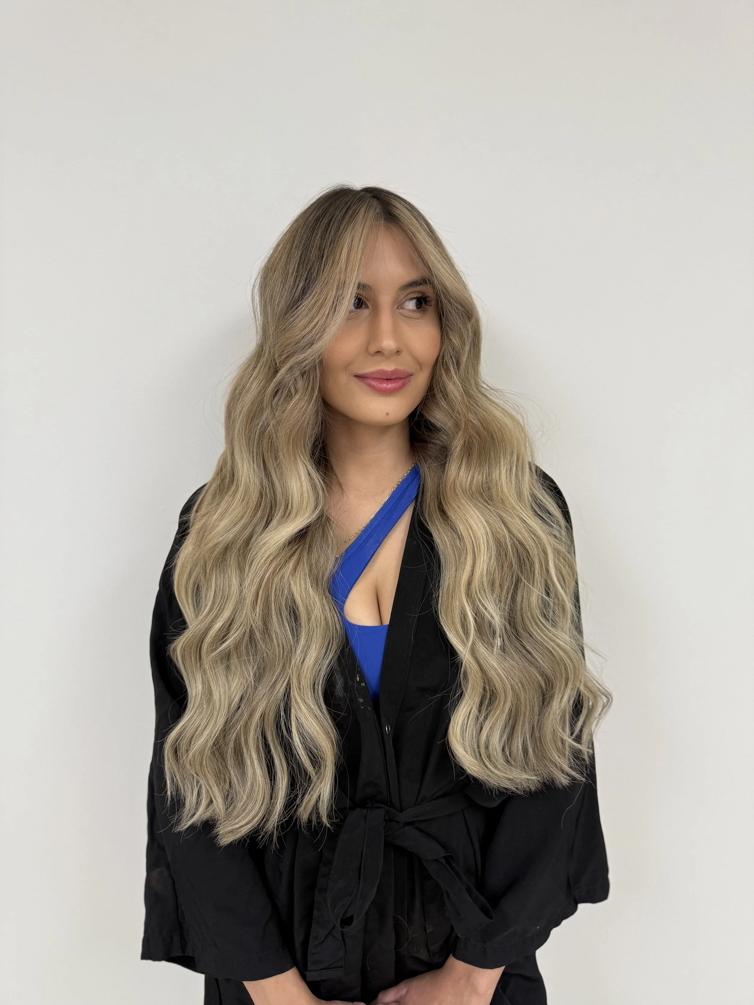 A woman with long, wavy blonde hair wearing a black jacket over a blue top, standing against a plain white wall.