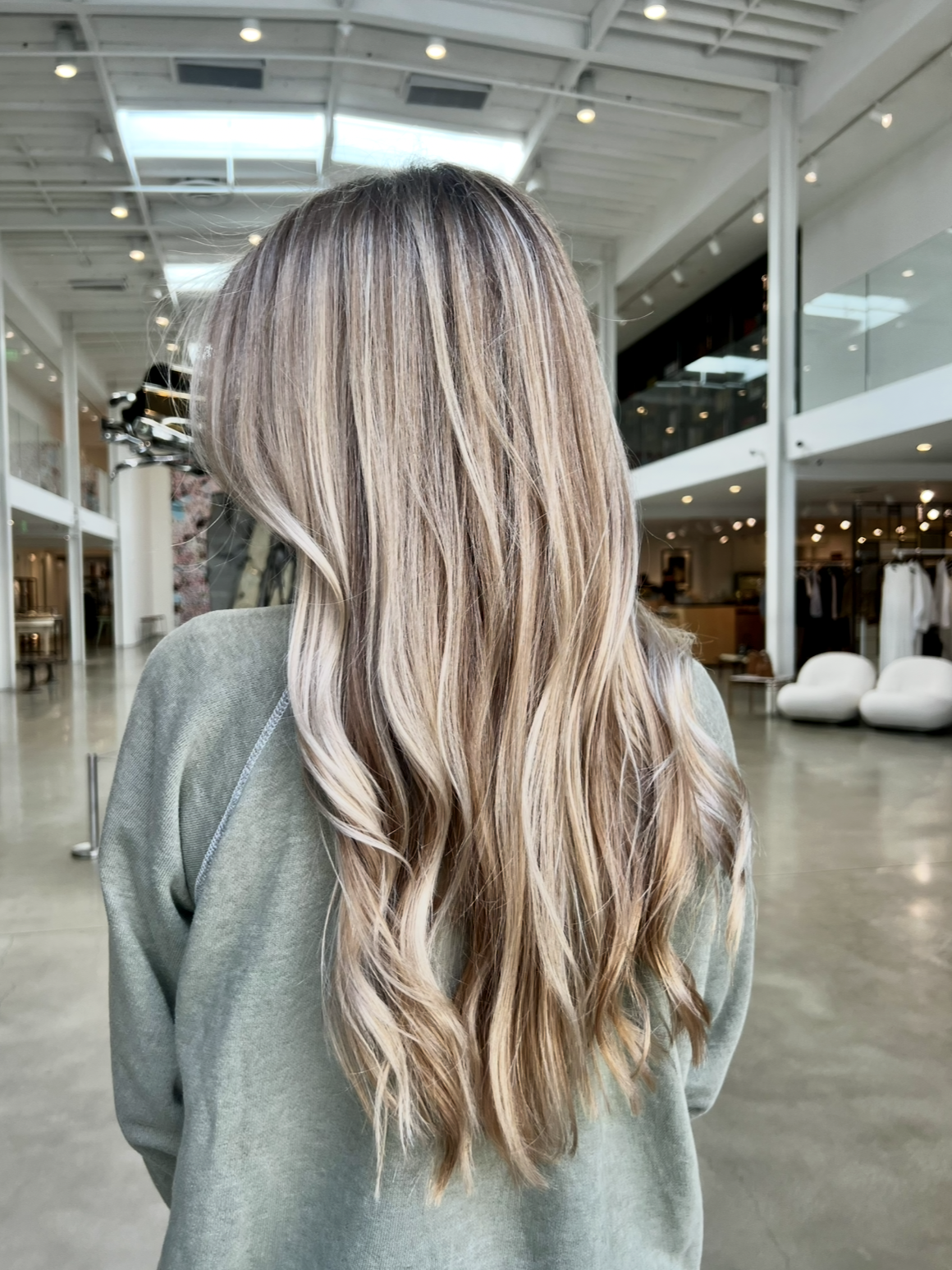 Back view of a woman with long, wavy, light brown hair with blonde highlights inside a modern, open-plan building with a high glass ceiling and white furniture.