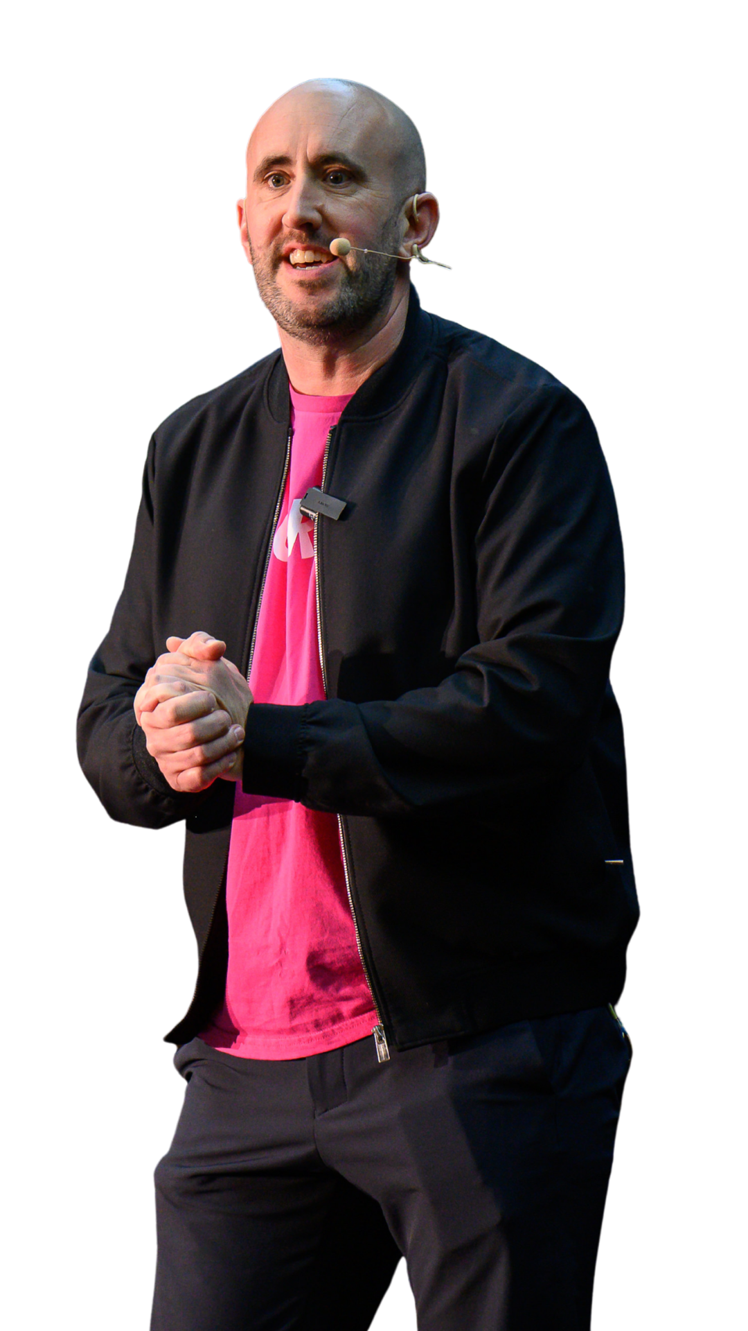 A man speaking at a conference, wearing a black jacket and pink shirt, holding his hands together.