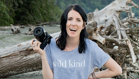 Woman holding a camera on a beach with driftwood and forest in the background, smiling
