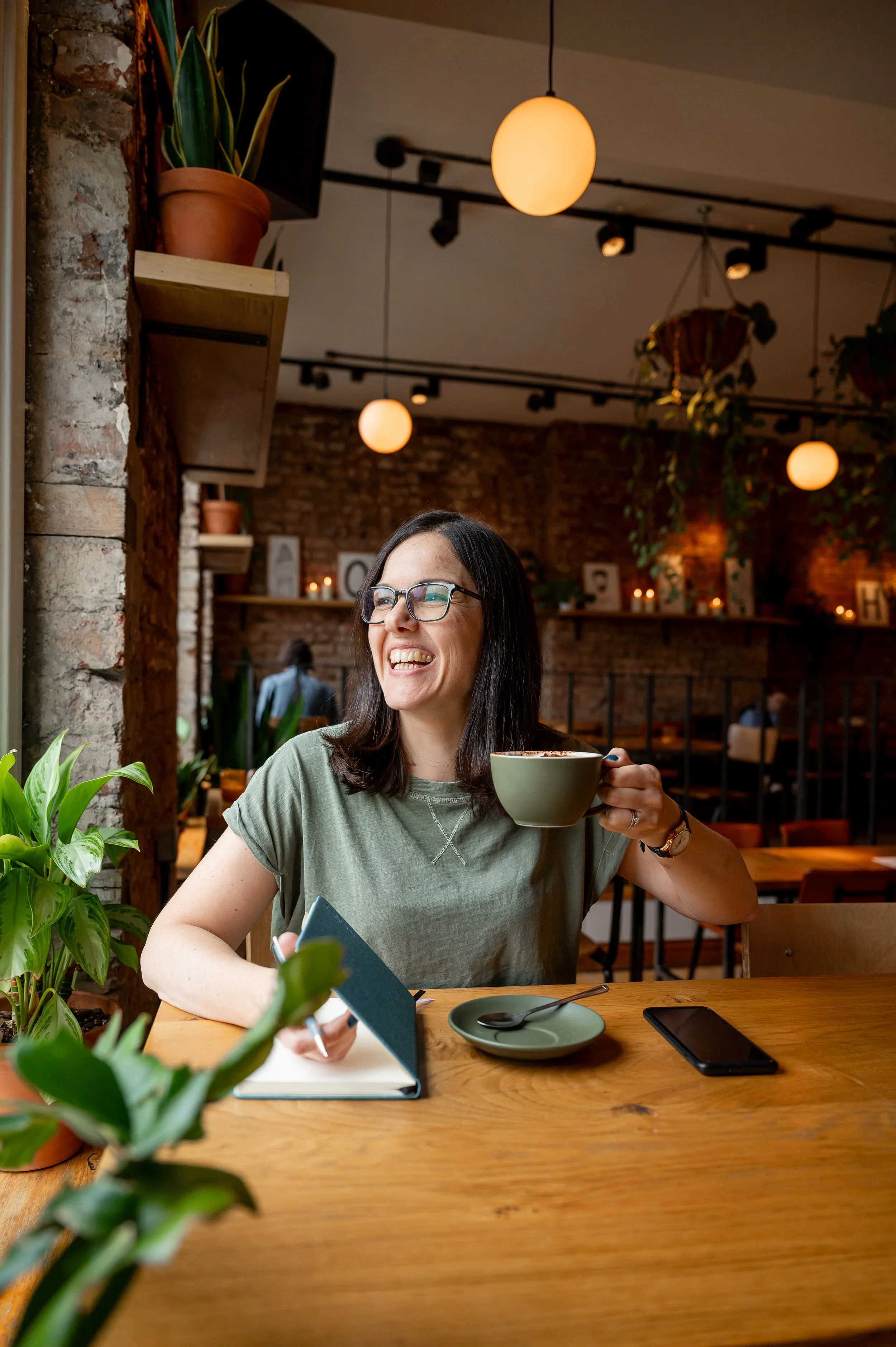 A woman with glasses and a green shirt smiling while sitting at a table, holding a cup of coffee. There is a notebook, a smartphone