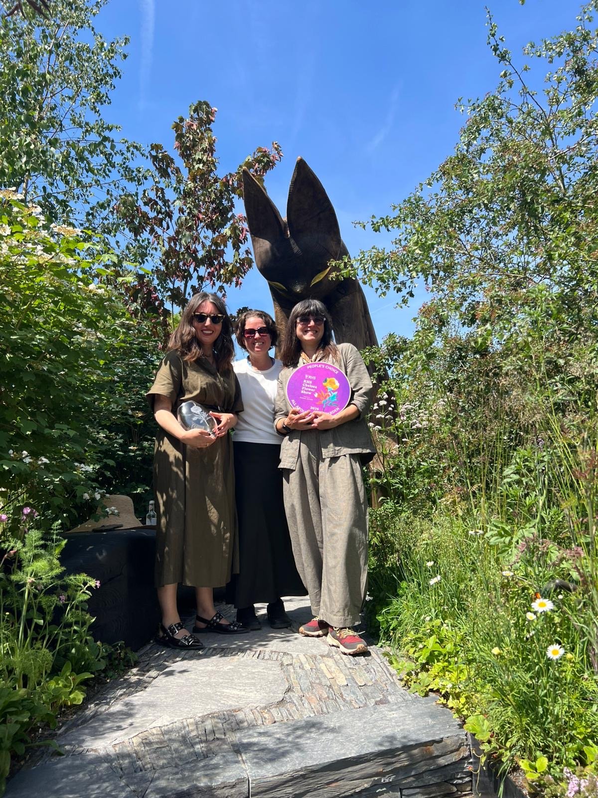 Three women standing outdoors in front of a large bat statue, holding awards and a colorful plaque, surrounded by lush green plants and trees, under a bright blue sky.