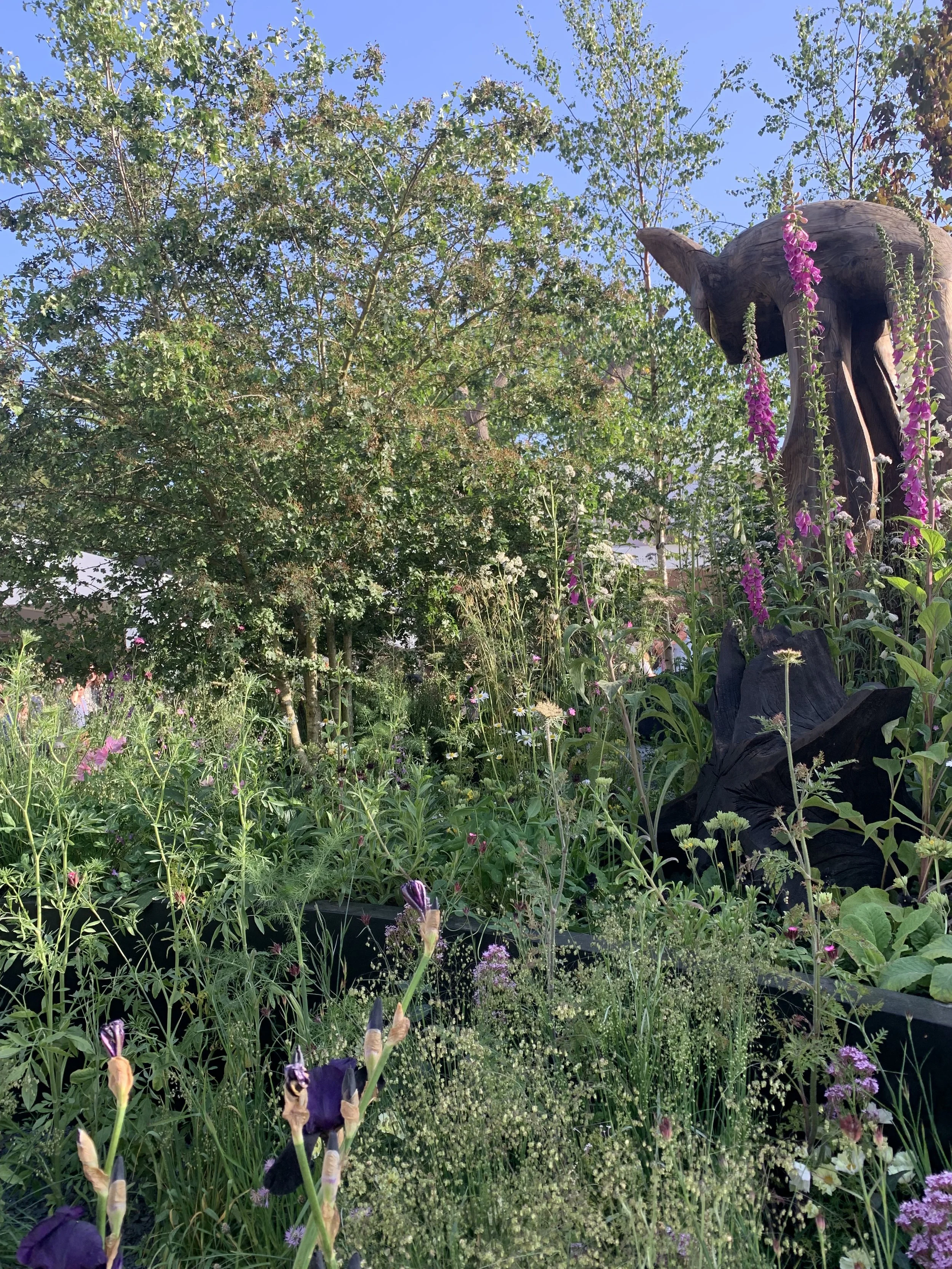 A lush garden scene with various flowers, trees, and a wooden sculpture of an bat, surrounded by colorful plants and greenery under a clear blue sky.