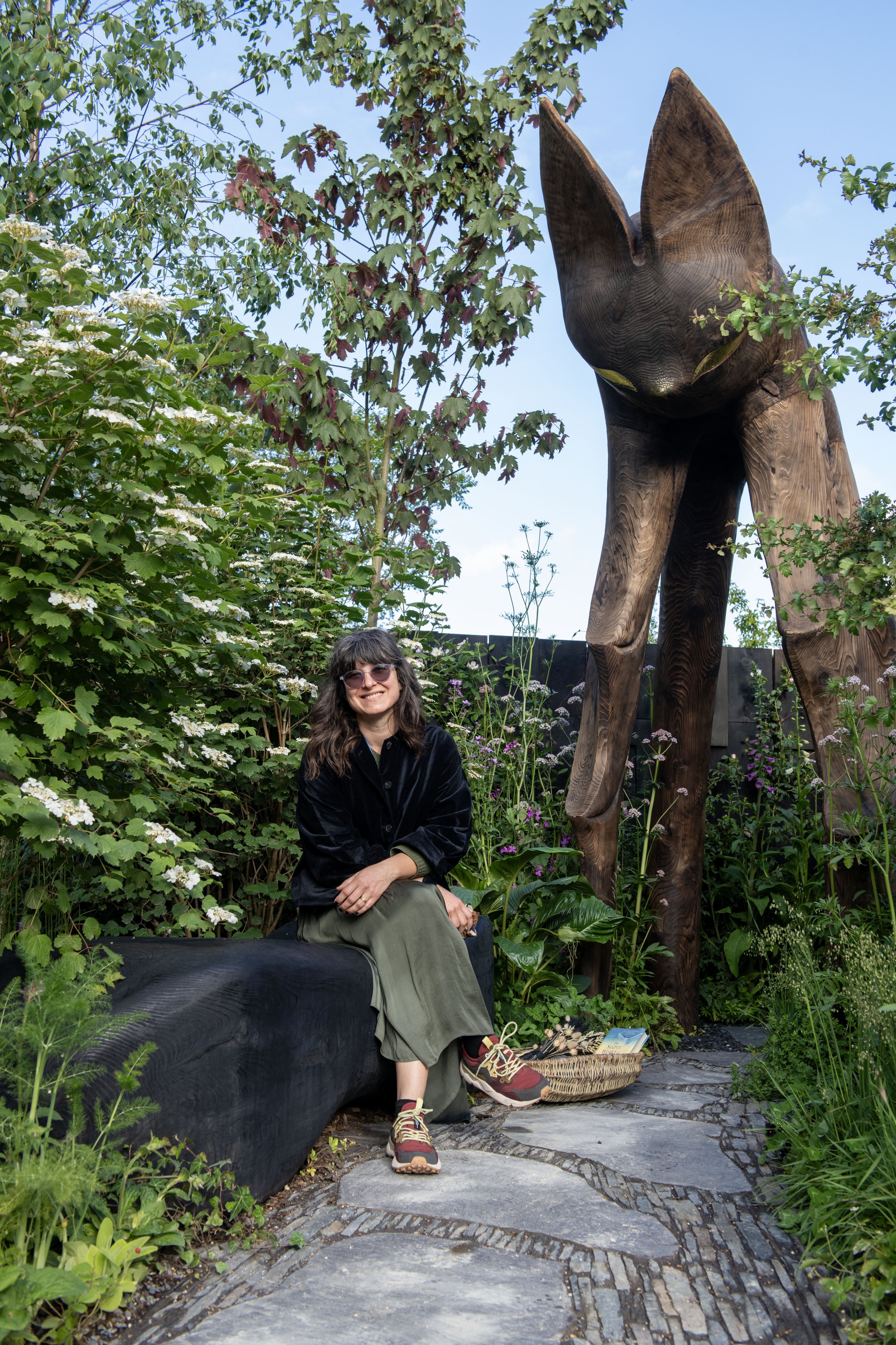 Cecilia is sitting on a bench in a lush garden with various green plants and flowers, beside a tall wooden bat sculpture.