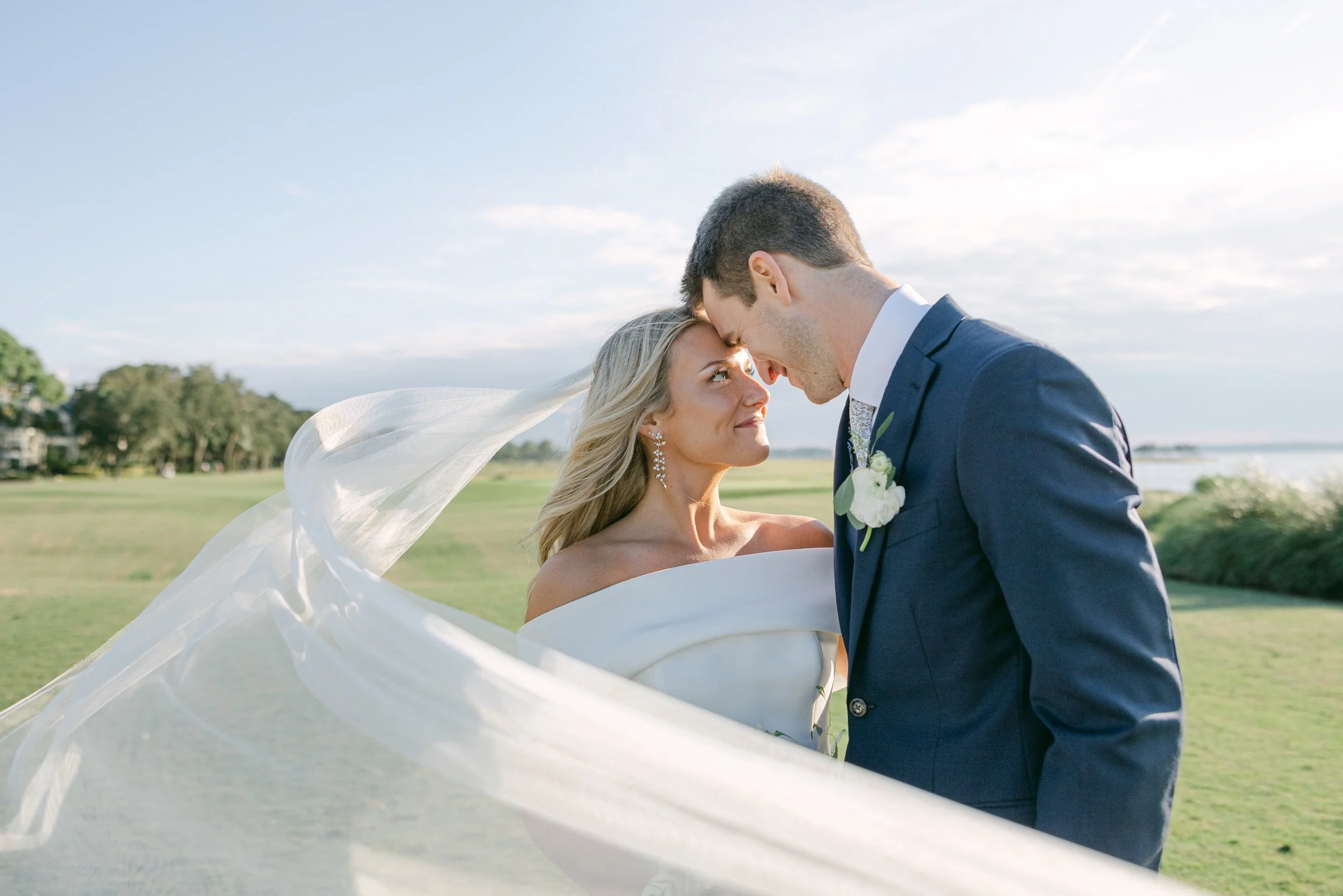 18th green Sea Pines Harbor Town. A bride and groom share a close moment outdoors, with the bride in a white dress and veil and the groom in a dark suit, in a scenic grassy area with trees and a cloudy sky in the background.