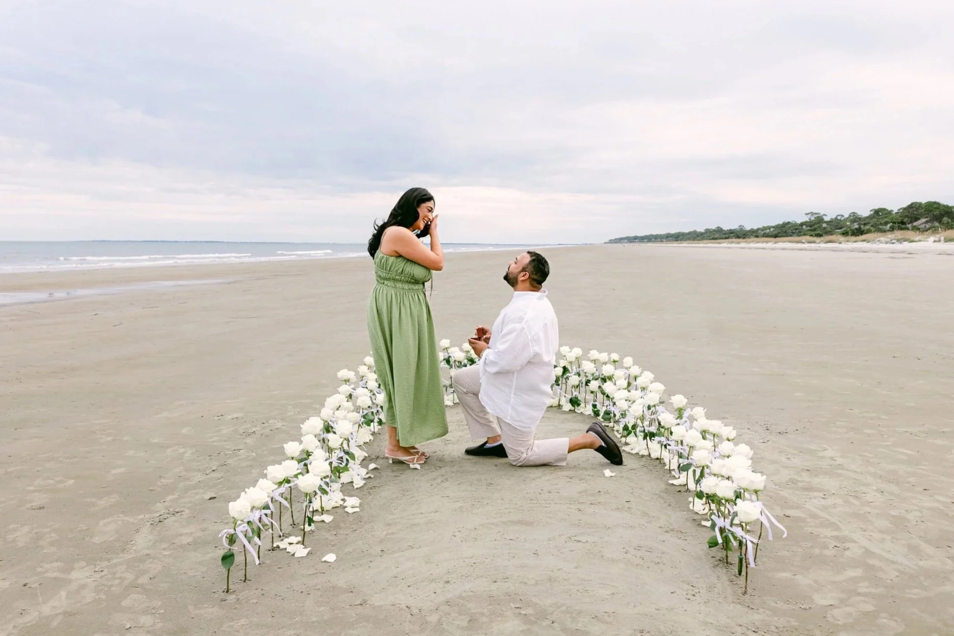 A couple sharing a marriage proposal on a beach, with the man kneeling on one knee surrounded by white flowers, while the woman appears happily surprised.