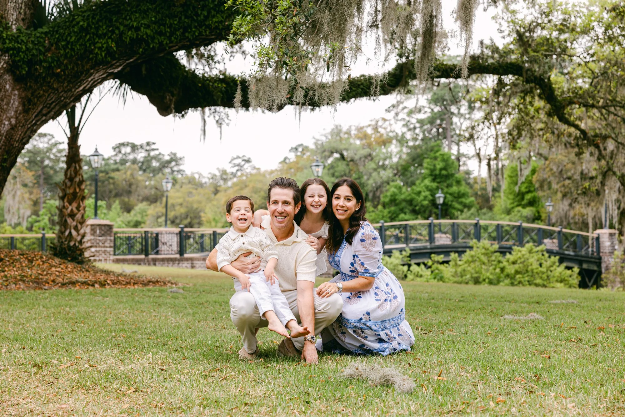 Family of four smiling and posing outdoors under a large moss-covered tree in a park with a small bridge in the background.