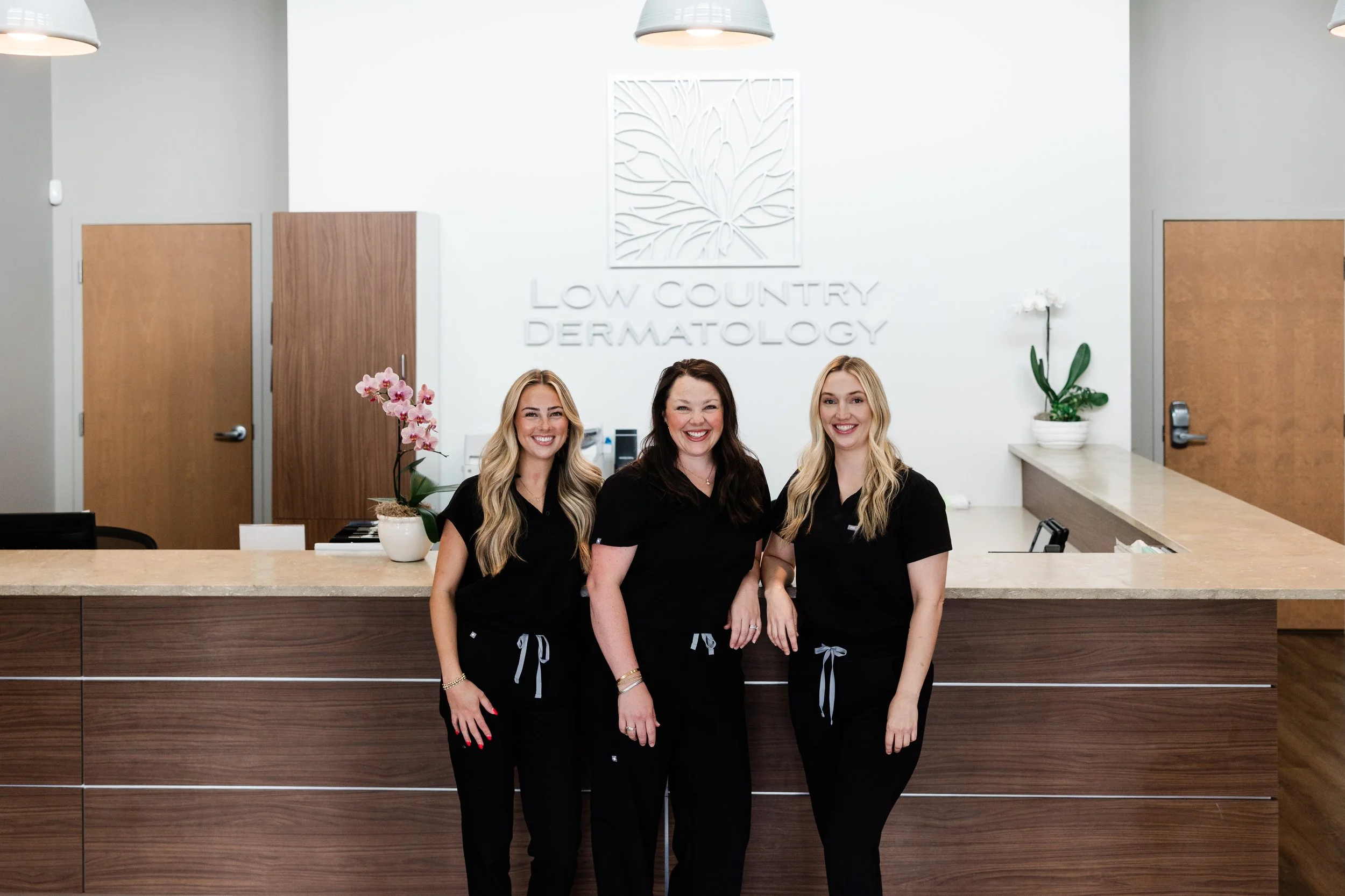 Three smiling women in black uniforms standing behind a reception desk in a dermatology clinic with plants and the clinic's name, Low Country Dermatology, on the wall behind them.