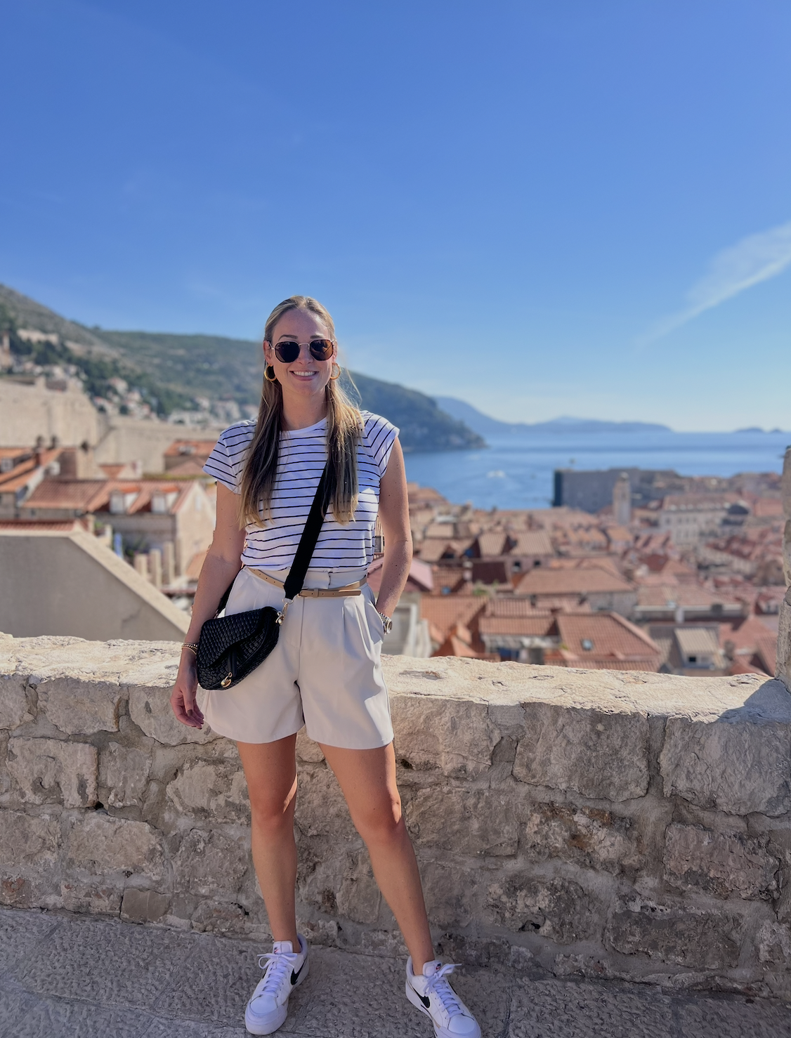A young woman standing outdoors on a stone wall, smiling, with a scenic view of rooftops, a body of water, and hills in the background. She is wearing sunglasses, a striped t-shirt, beige shorts, and sneakers, carrying a black crossbody bag.