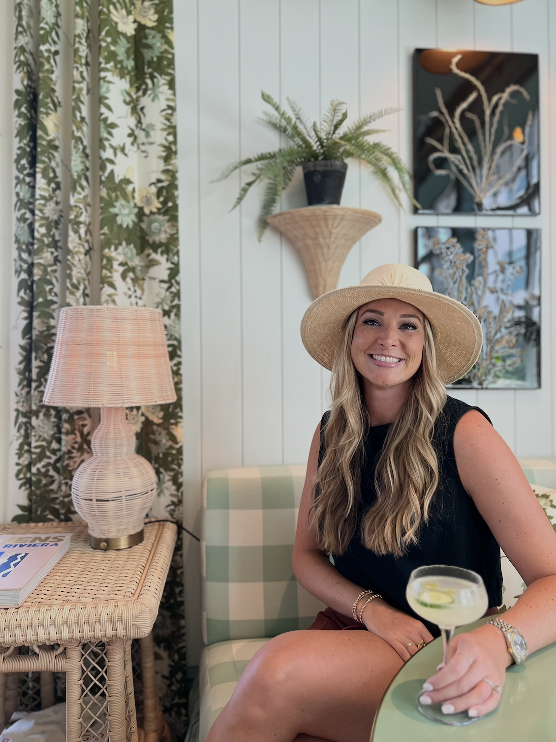 Red Letter Travel advisor, Ansley Robertson, wearing a straw sun hat and a black sleeveless top, sitting on a green and white checkered couch in a tropical-themed room, holding a glass of cocktail with lime slices and looking at the camera, smiling.