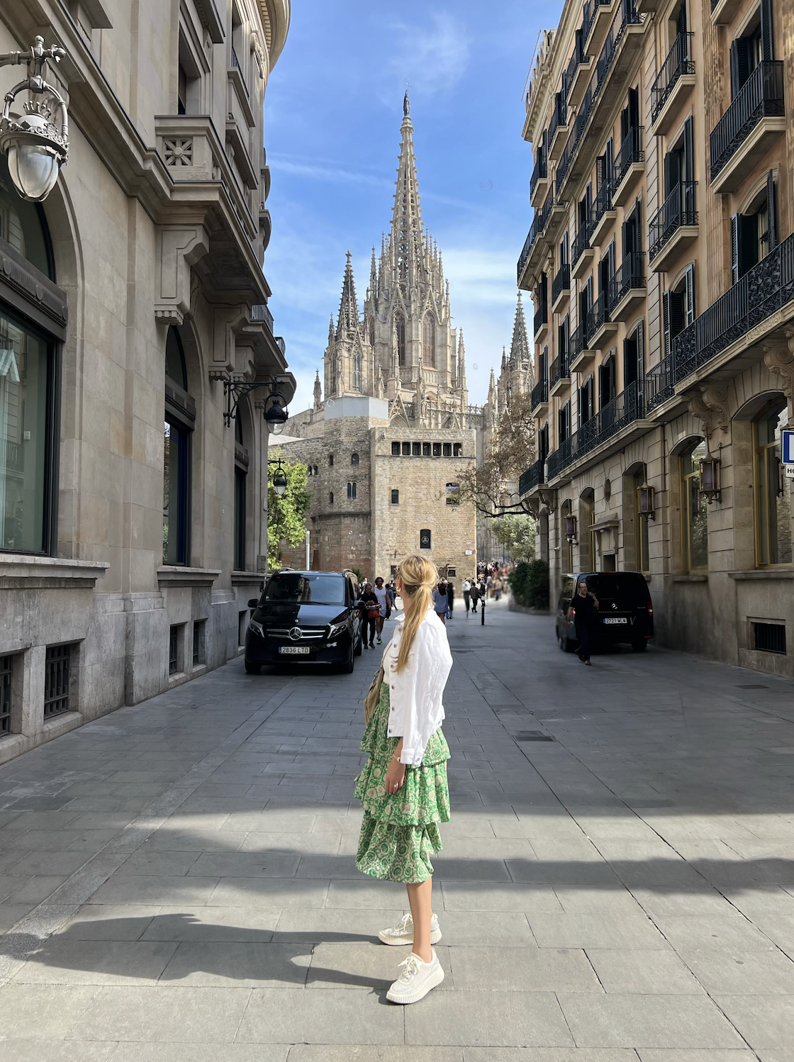 A woman with long blonde hair in a green patterned dress, white jacket, and sneakers standing on a city street, with the Gothic-style Cathedral of Barcelona in the background, surrounded by older buildings and parked cars.