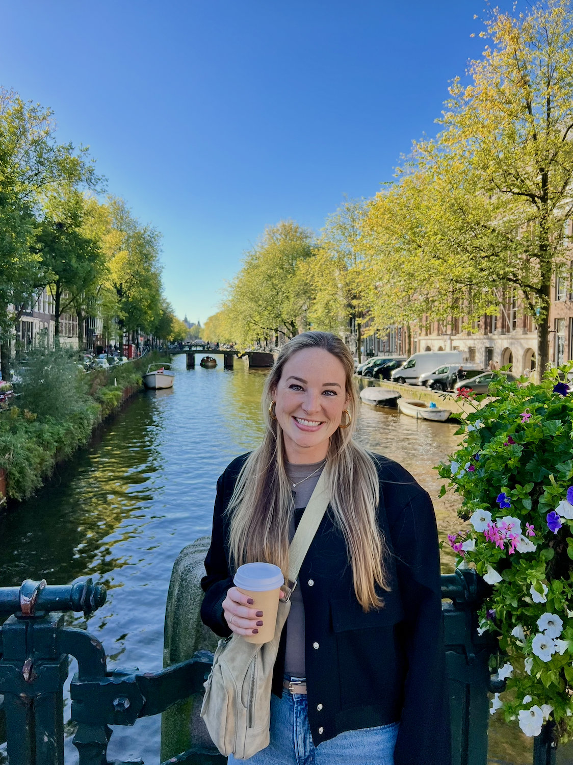 A woman smiling holding a coffee cup standing near a canal lined with trees and buildings, with boats on the water and a bridge in the background, on a bright, sunny day.