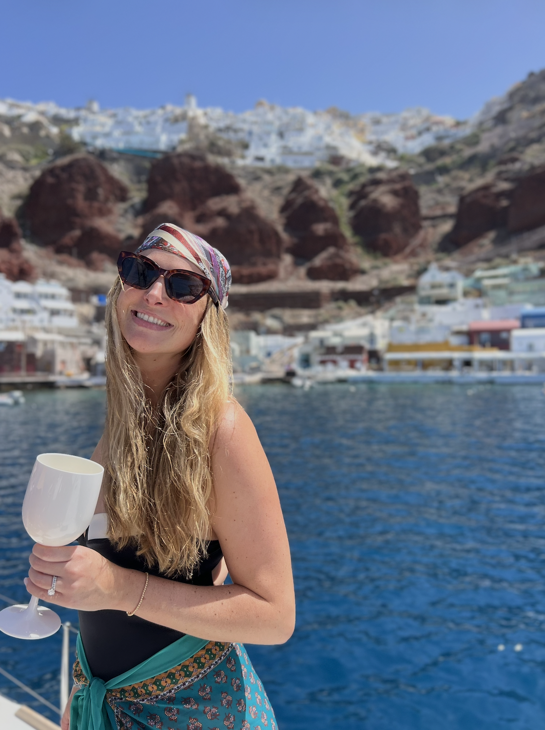 Ansley Robertson, a Red Letter Travel advisor, wearing sunglasses and a bandana, holding a white wine glass, standing near a body of water with a coastal village and hillside with white buildings in the background.