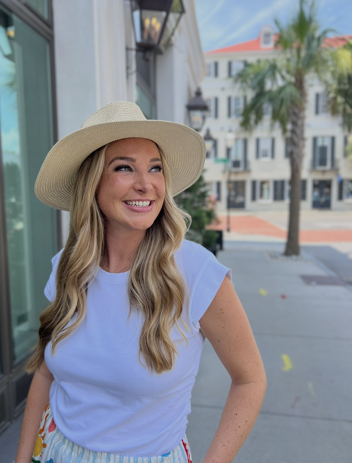 Ansley Robertson in Charleston wearing a wide-brimmed straw hat and a white t-shirt, smiling outdoors on a sunny day in an urban area with palm trees and multi-story buildings in the background.