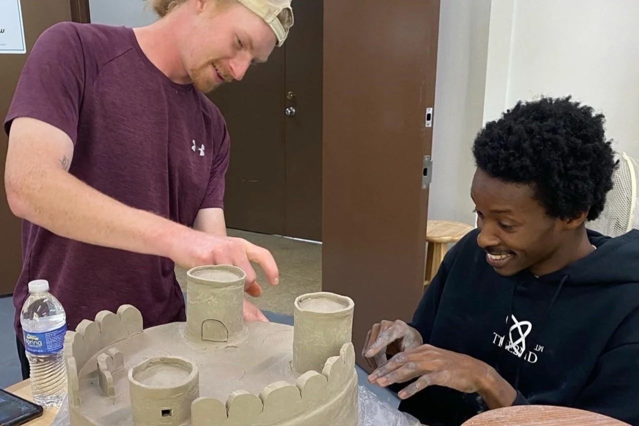 Student & teacher working together on a clay project, creating a small castle with towers and walls, in an indoor setting.
