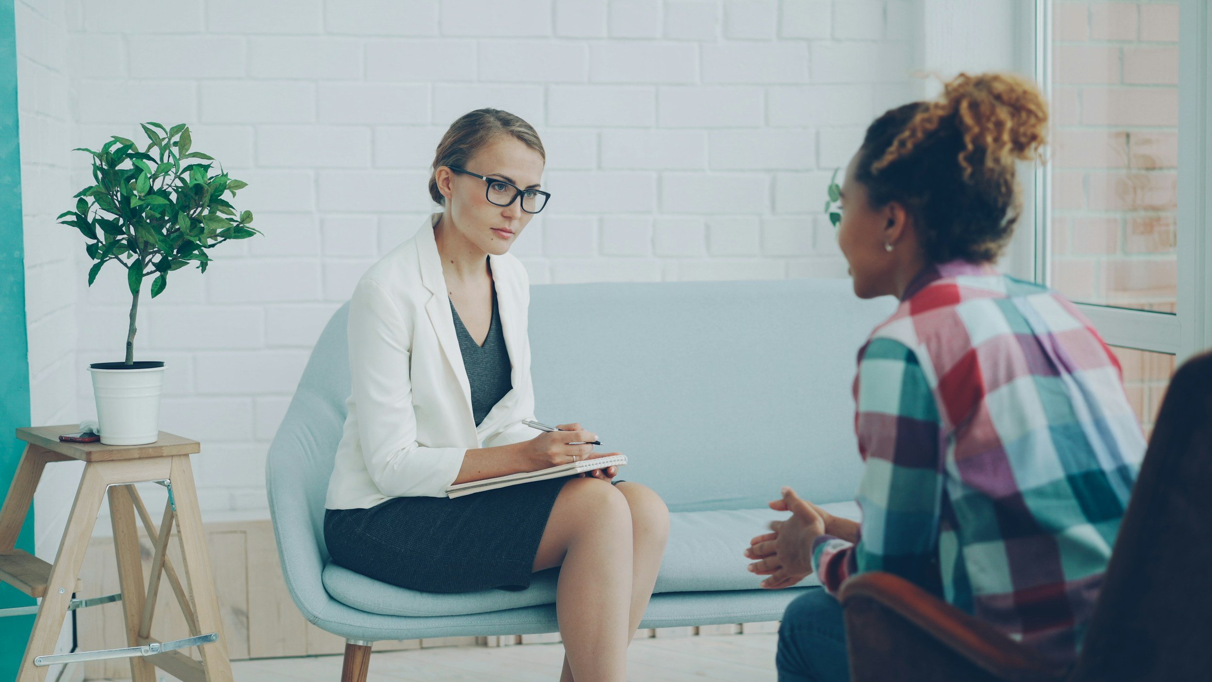 A woman with short brown hair, glasses, and a white blazer taking notes on a notepad while talking to a woman with curly hair wearing a colorful plaid shirt, sitting in a modern, bright room with a potted plant on a small wooden table.
