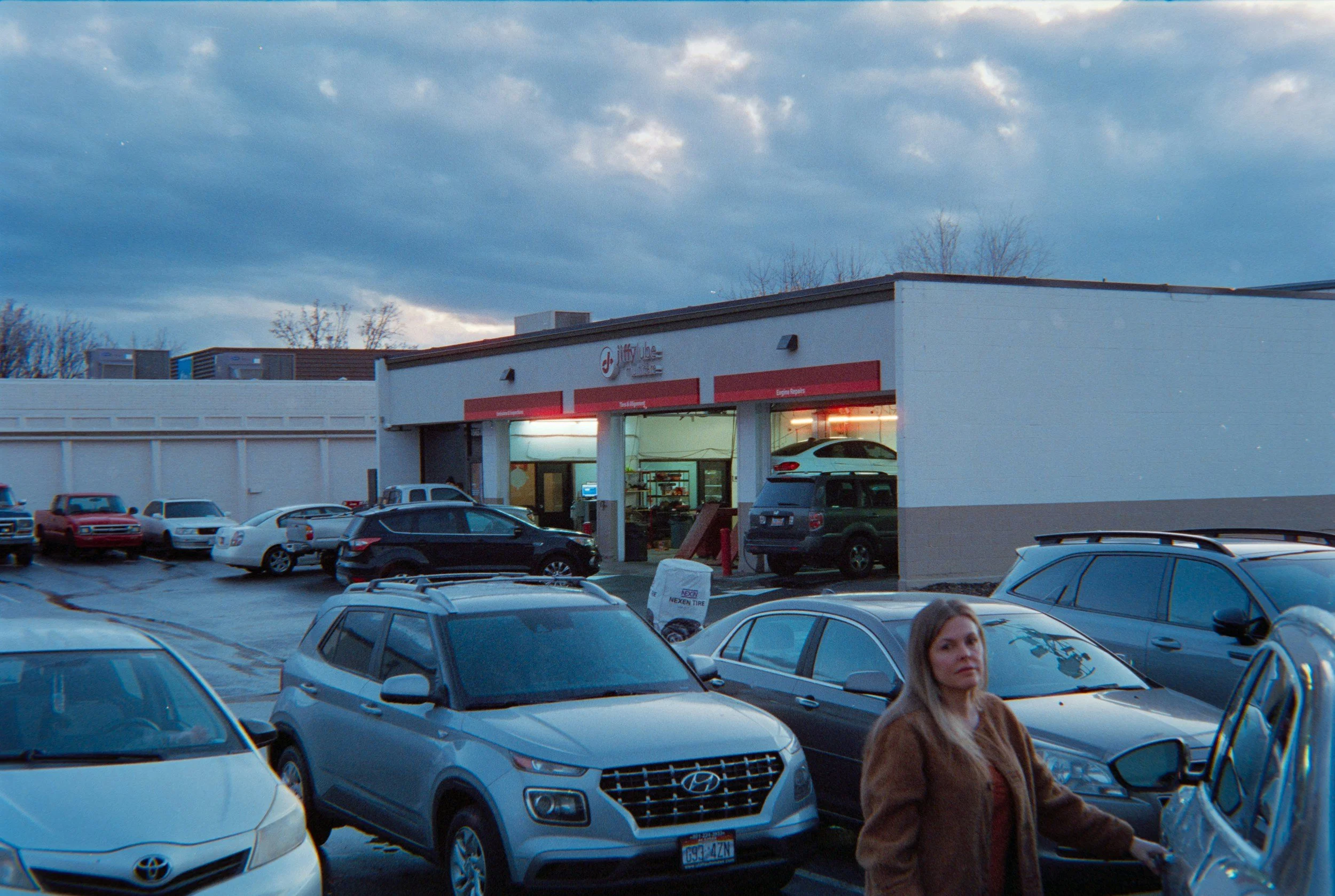 A parking lot in front of a white auto repair shop with cars parked and a woman walking. The shop has a red sign with white lettering, and the sky above is partly cloudy.