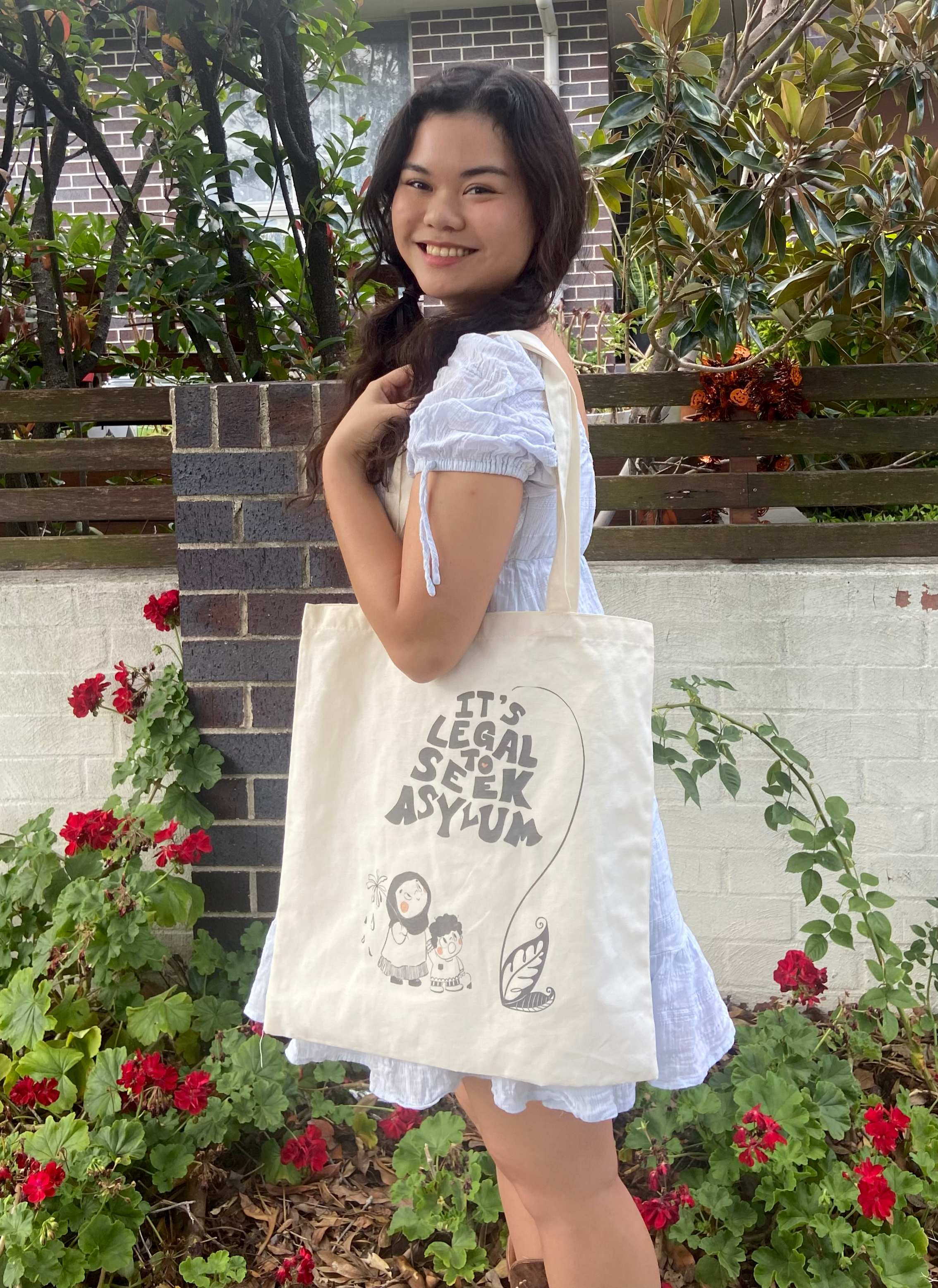 Artist Audrey Villafaña standing outside, carrying a tote bag with an illustration of two people and the words "it's legal to seek asylum" in the shape of a bluebell flower.