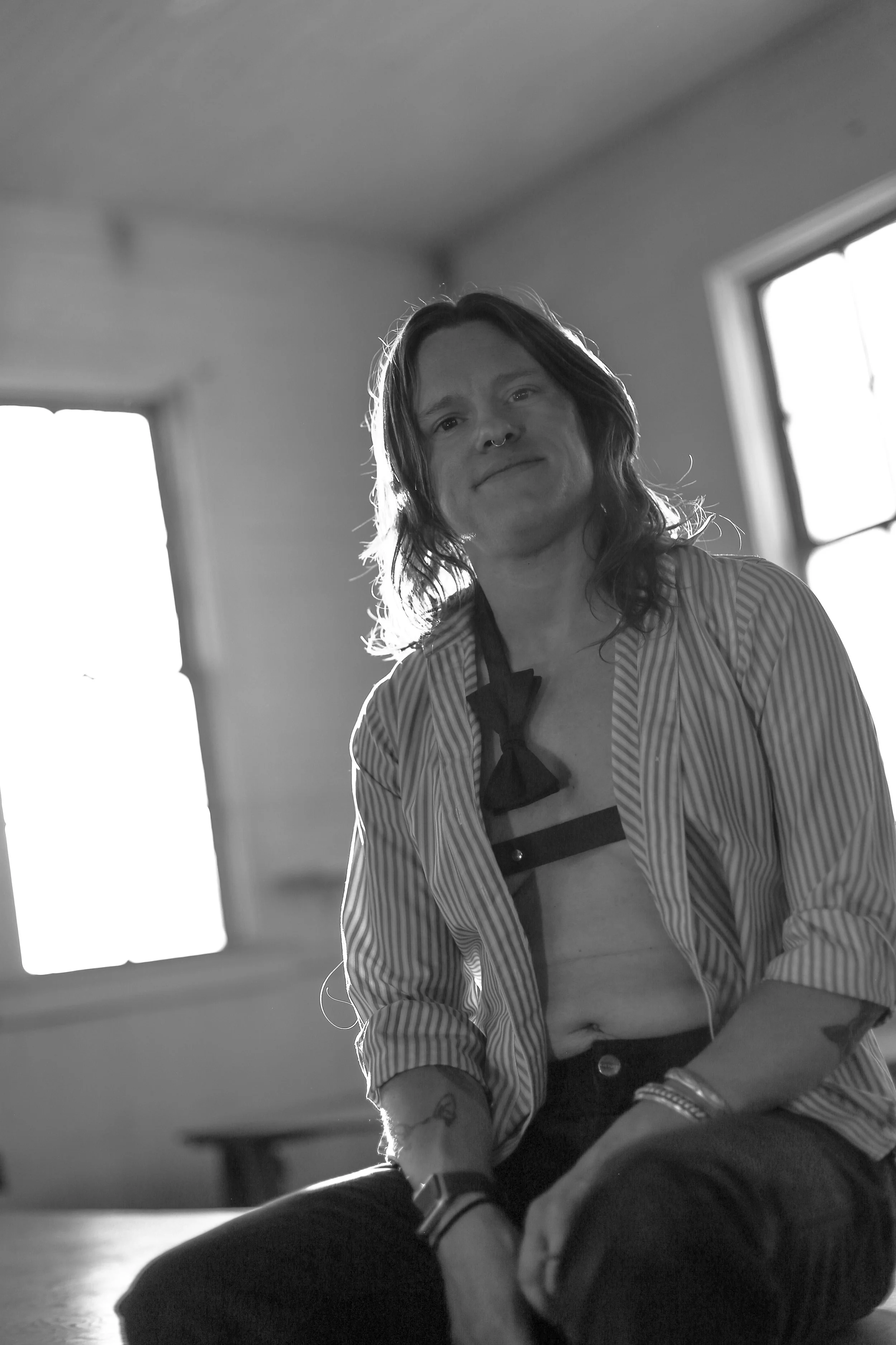 A certified sex coach for queer and trans people and couples sits smiling in a room with bright windows. They are trauma-informed, a certified sex and relationship coach, and help with lack of orgasm or less orgasms