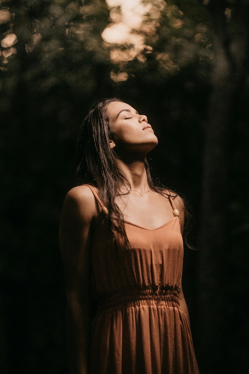 A woman standing outdoors with eyes closed and face tilted upward wearing a brown dress, representing surrender, release, and embodied healing