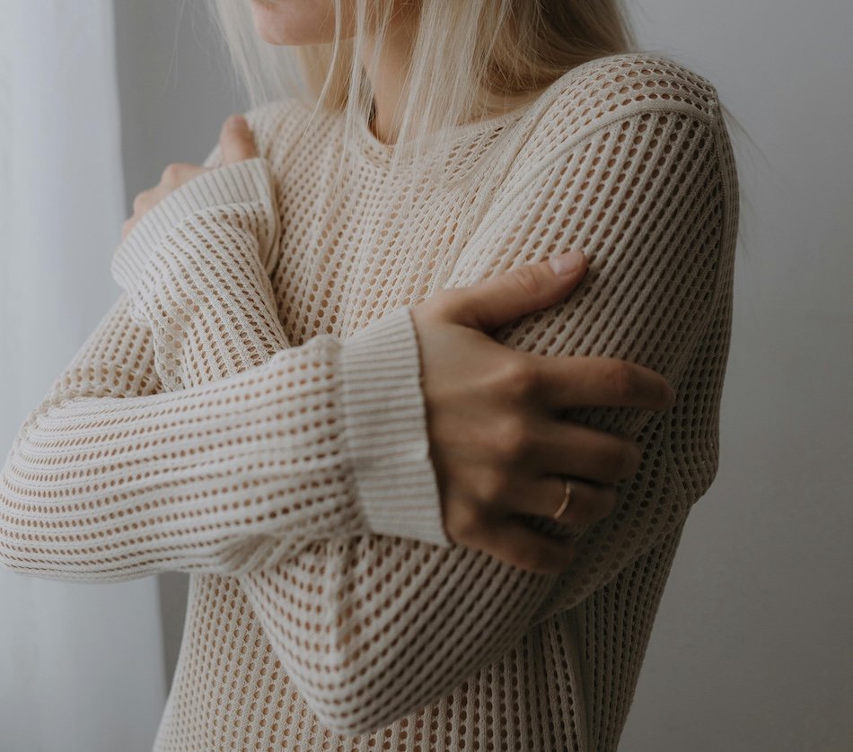A woman in a beige knit sweater with arms crossed and hands resting on her shoulders, representing self-soothing and nervous system regulation