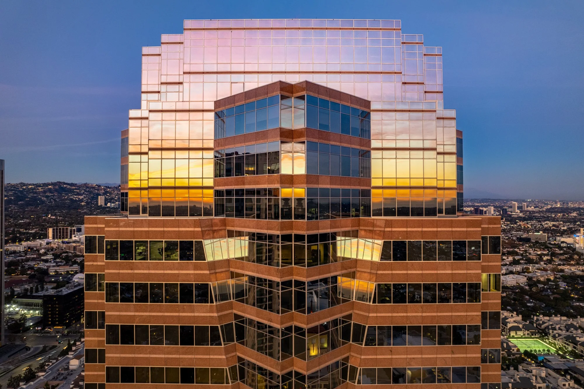 A tall modern office building with reflective glass windows during sunset, with a sunset sky reflected on the windows and a cityscape with hills in the background.
