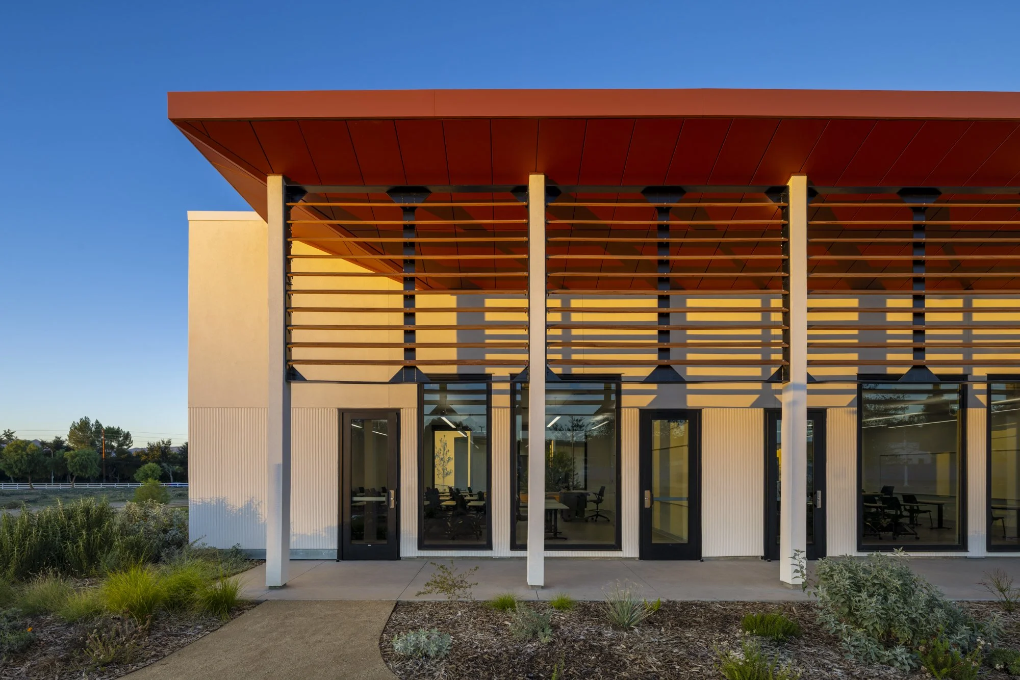 Modern rectangular building with wooden exterior and large glass window, surrounded by yellowish plants, mountains in the background.