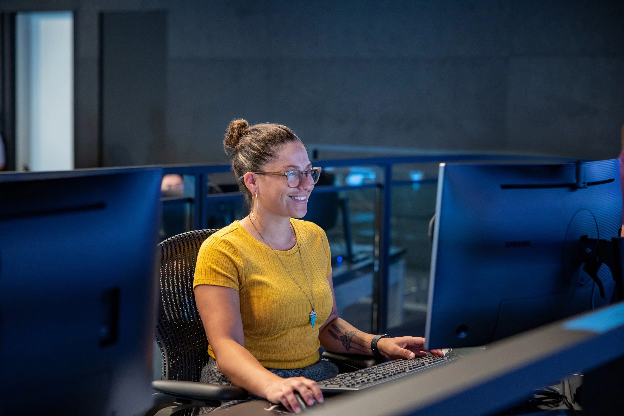 A woman with glasses and a bun, wearing a yellow shirt, sitting at a desk and working on a computer in an office environment.