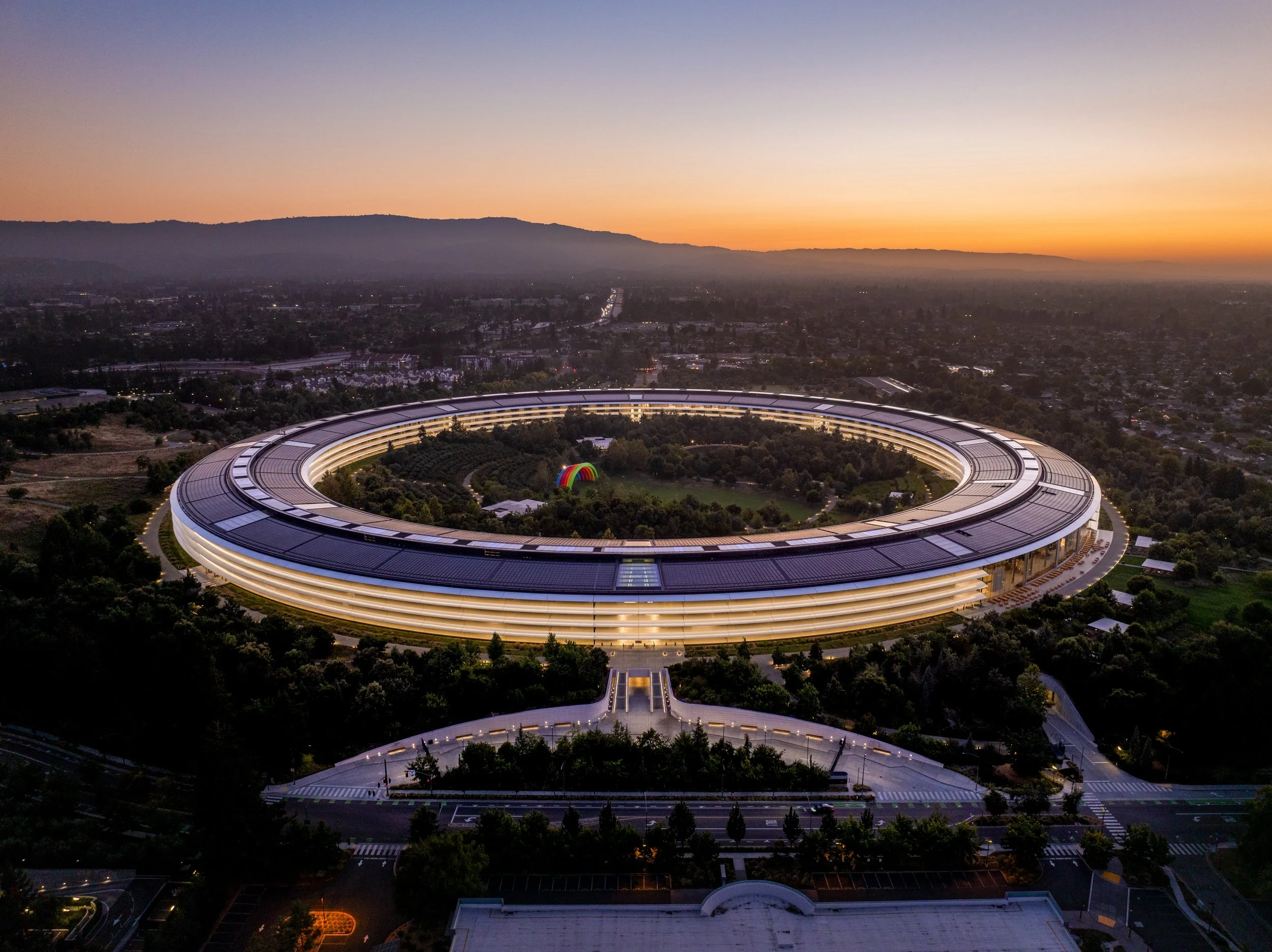 An aerial view of a circular building with a roof lined with solar panels, surrounded by trees and cityscape, at sunset with mountain range in the background.