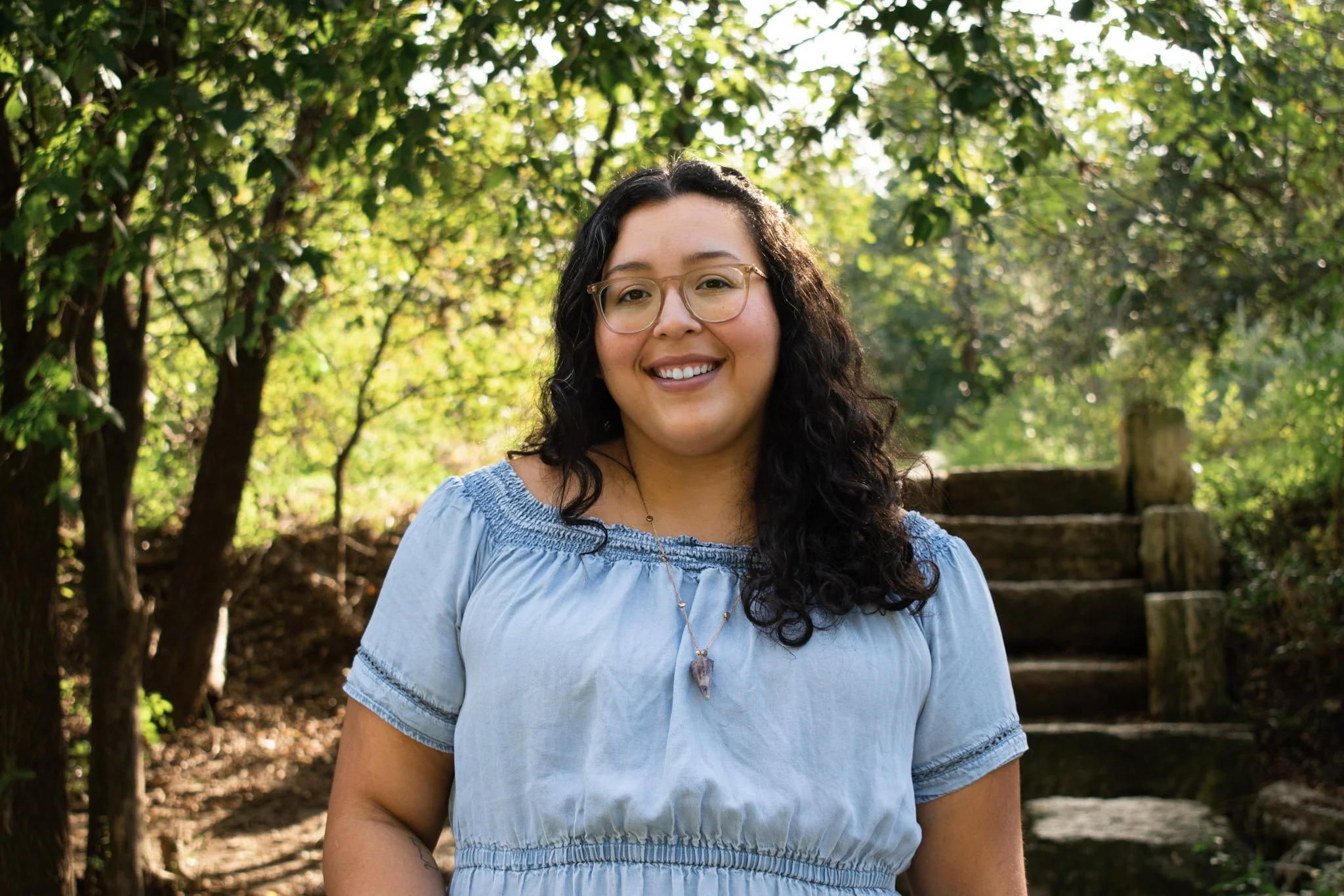 Courtney smiles at the camera. She has medium length curly black hair, brown glasses, & wears a denim blue dress with a pendant necklace. Green trees are in background.