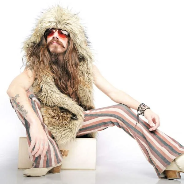 Man with long hair and beard wearing a fur vest, striped bell-bottom pants, sunglasses, and multiple bracelets, sitting on a white block against a white background.