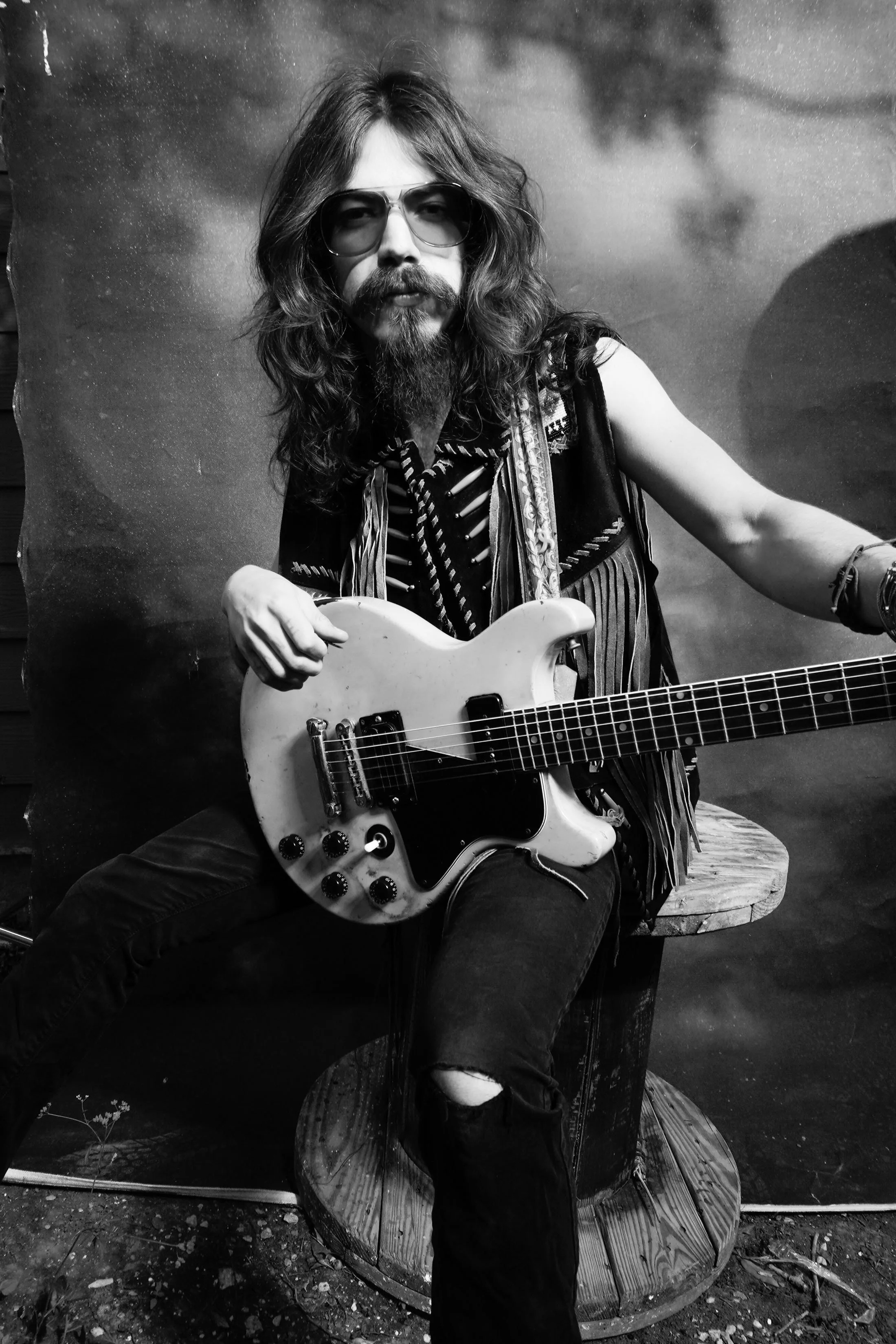 Black and white photo of a man with long hair, a mustache, and a beard, wearing sunglasses and a vest, sitting on a wooden spool, holding an electric guitar.