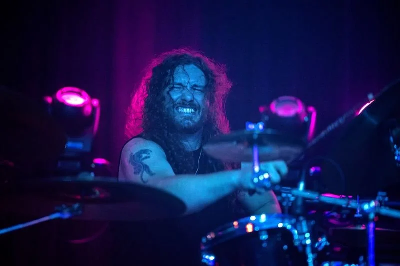 A drummer with long curly hair, a beard, and a tattoo on his arm playing drums on stage, smiling in front of purple stage lights.