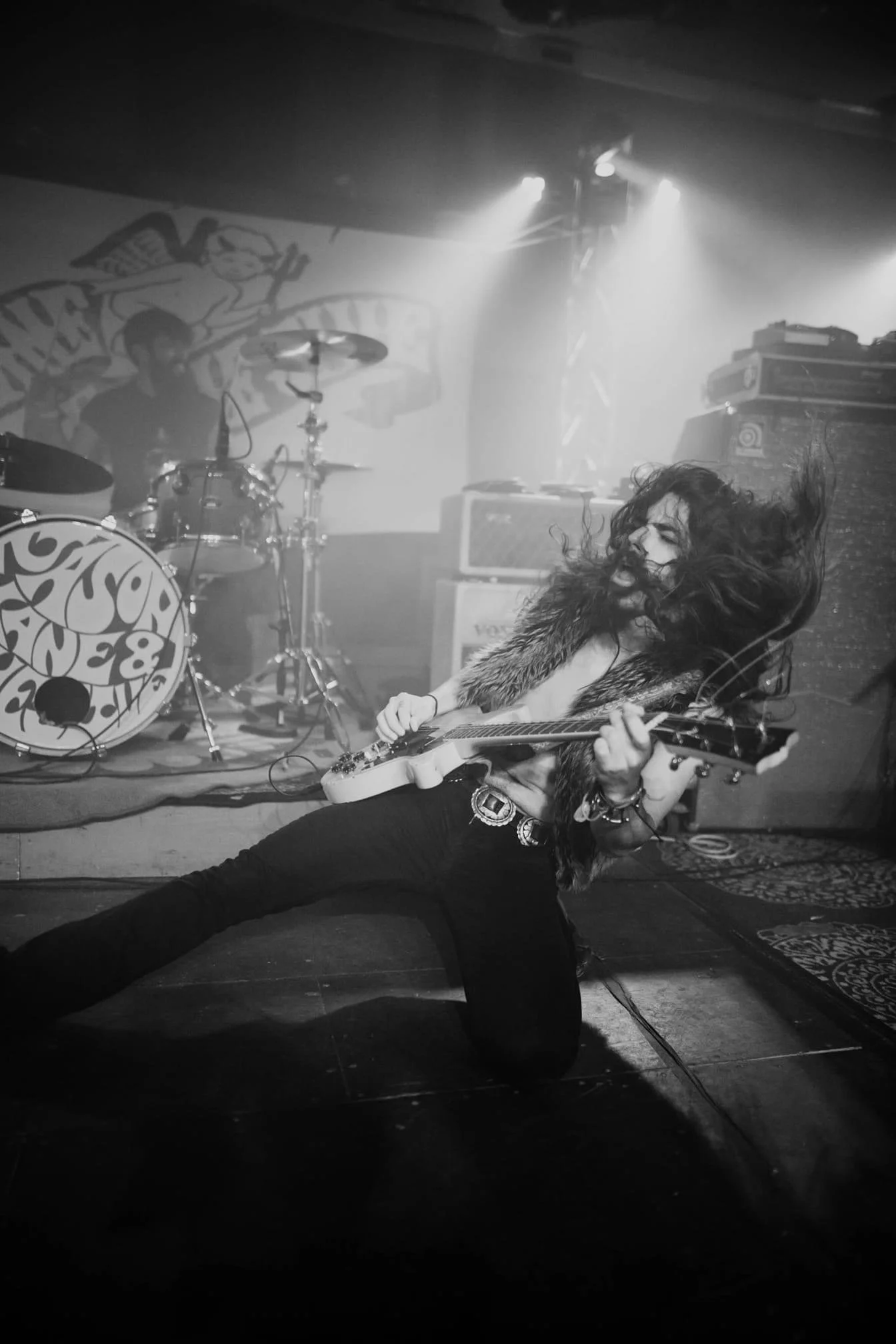 A rock musician passionately playing an electric guitar on stage, with long hair and no shirt, in a black-and-white photo. Another musician is playing drums in the background, with stage lights and sound equipment visible.