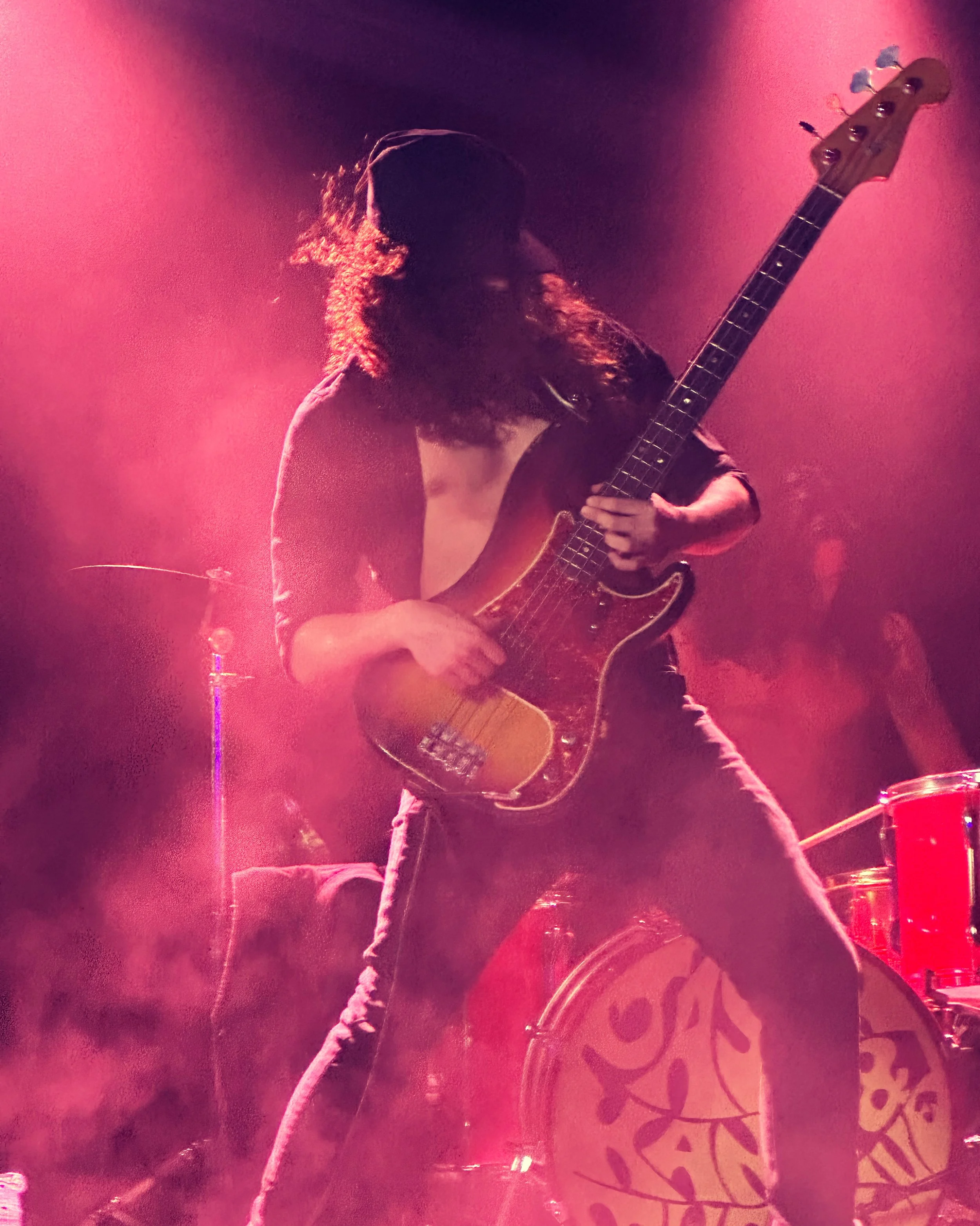 A musician with long hair playing an electric bass guitar on stage, illuminated by pink stage lights, with musical instruments and audience in the background.