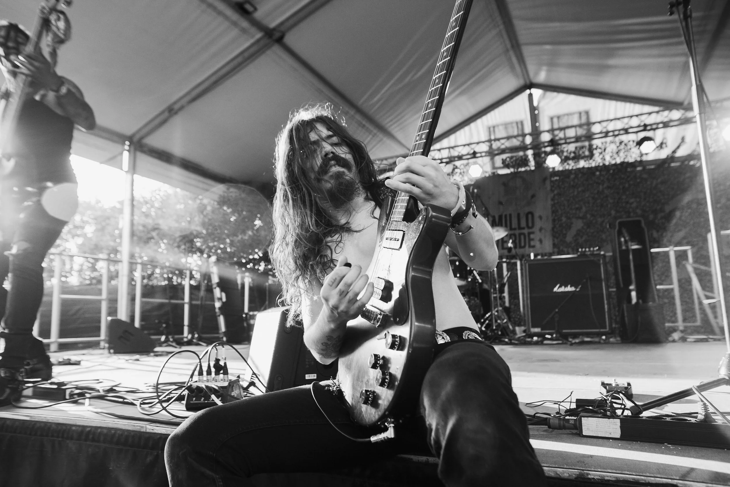A long-haired musician playing an electric guitar on stage during a live performance in an outdoor venue, with stage equipment and another performer visible in the background.