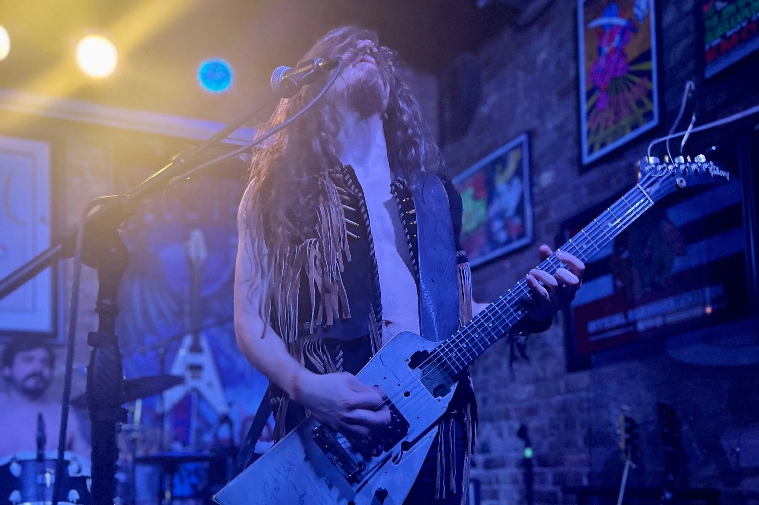 A musician with long hair, wearing a fringed vest, plays a guitar on stage next to a microphone in a dimly lit venue with colorful artworks on the wall.