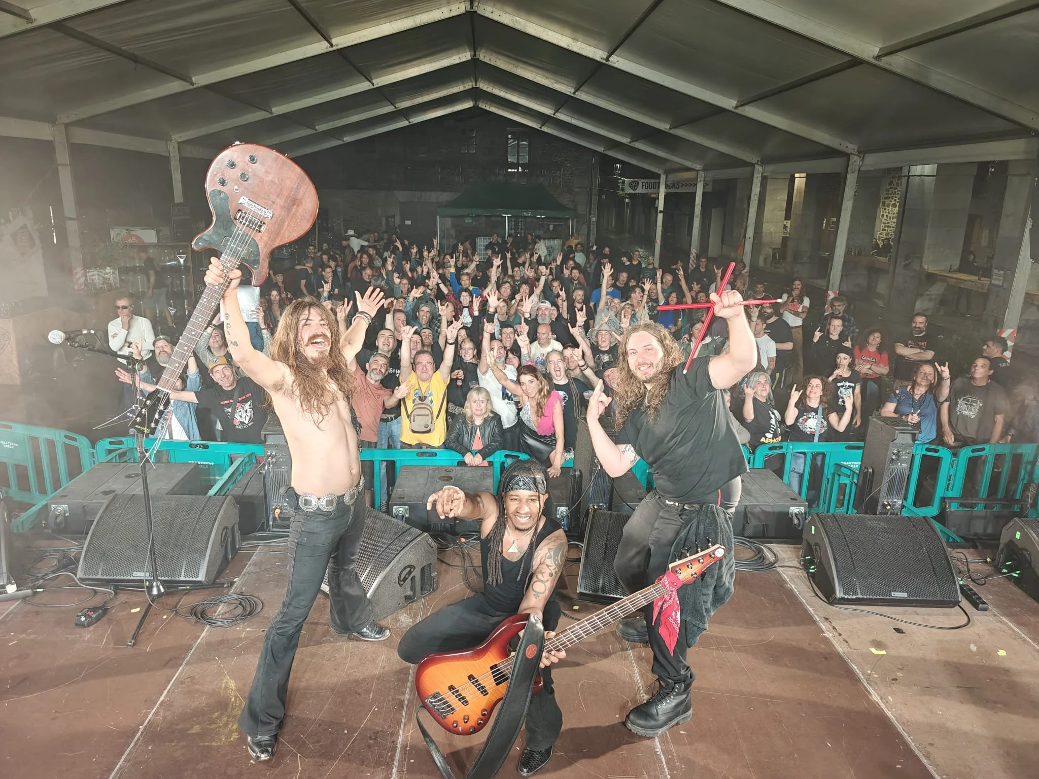 Three musicians on stage after a concert, with a crowd of fans in the background. The man on the left is shirtless, holding a bass guitar up high and smiling. The man in the middle is seated, holding an electric bass guitar, smiling, and wearing a bl