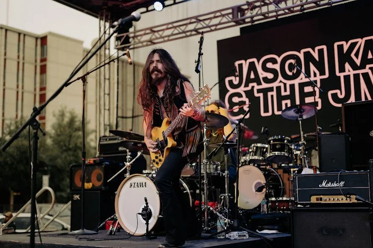 Musician with long hair and a beard playing an electric guitar on stage during a live outdoor concert.