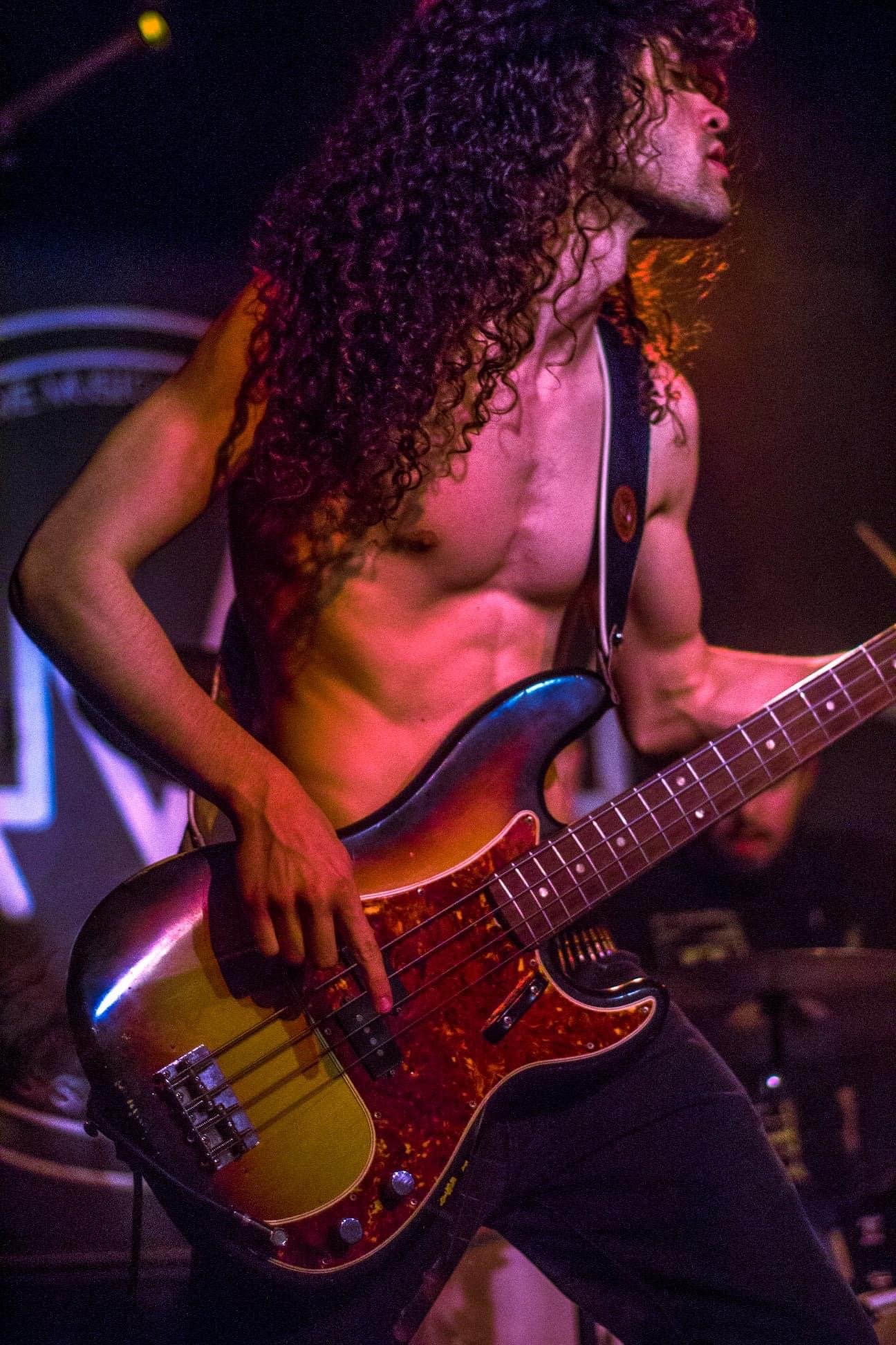 A shirtless male bassist with long, curly hair playing a bass guitar on stage.